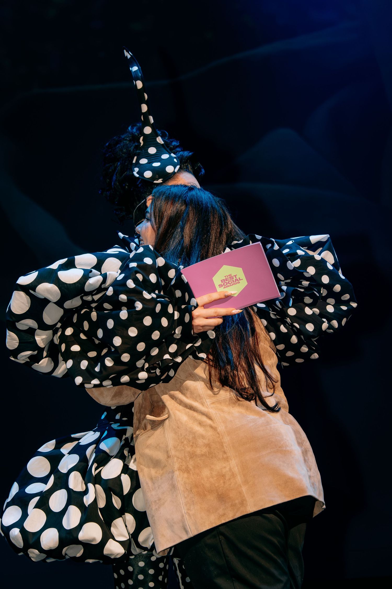 Performer in polka dot costume holding pink book on dark stage.