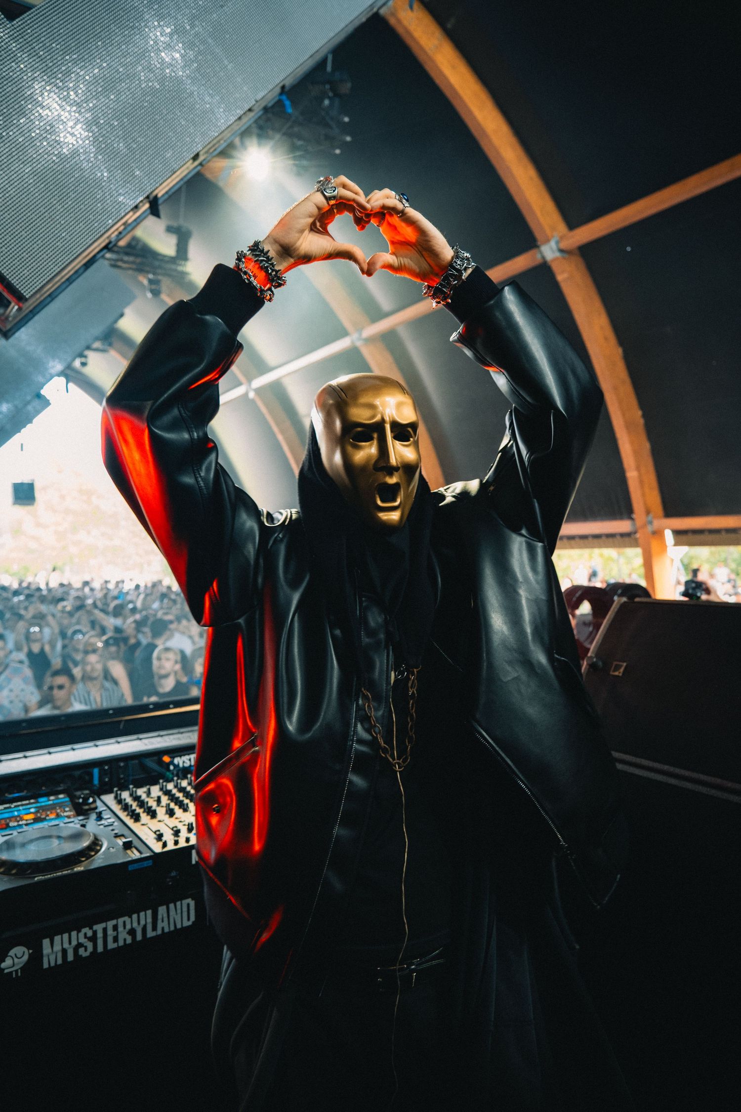 Masked DJ in black leather jacket making heart shape with hands above DJ booth at Mysteryland festival.