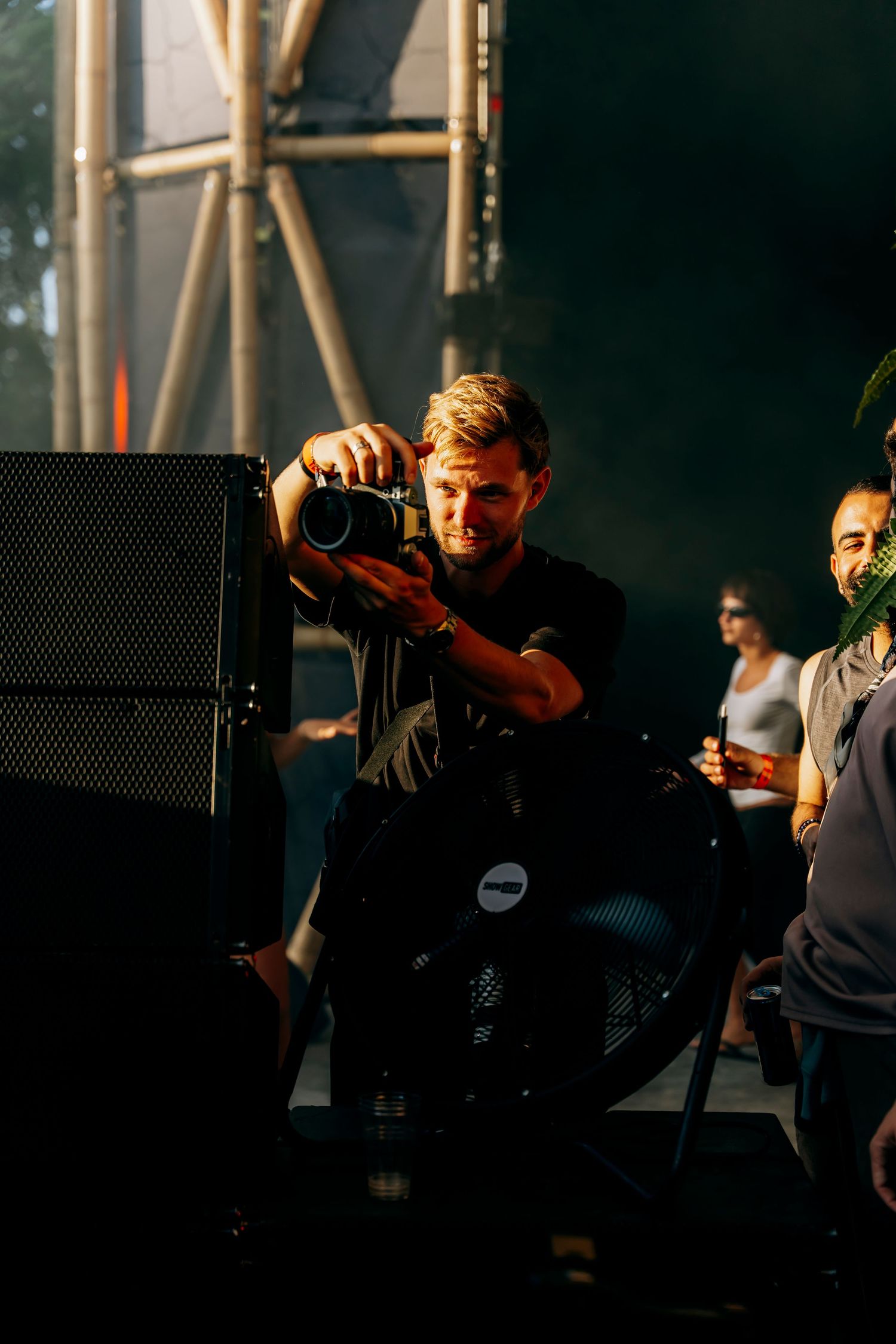 Blonde photographer crouching with camera at outdoor music festival stage.