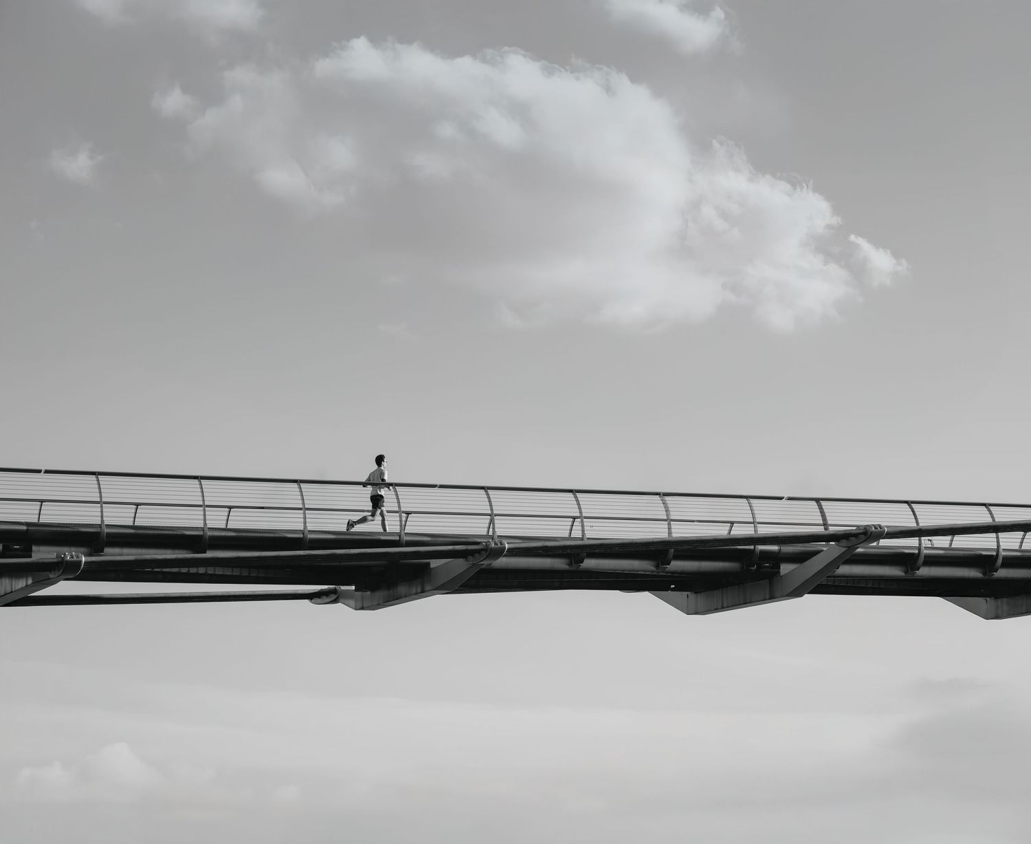 Person walking alone on a modern elevated bridge against a dramatic cloudy sky in black and white.