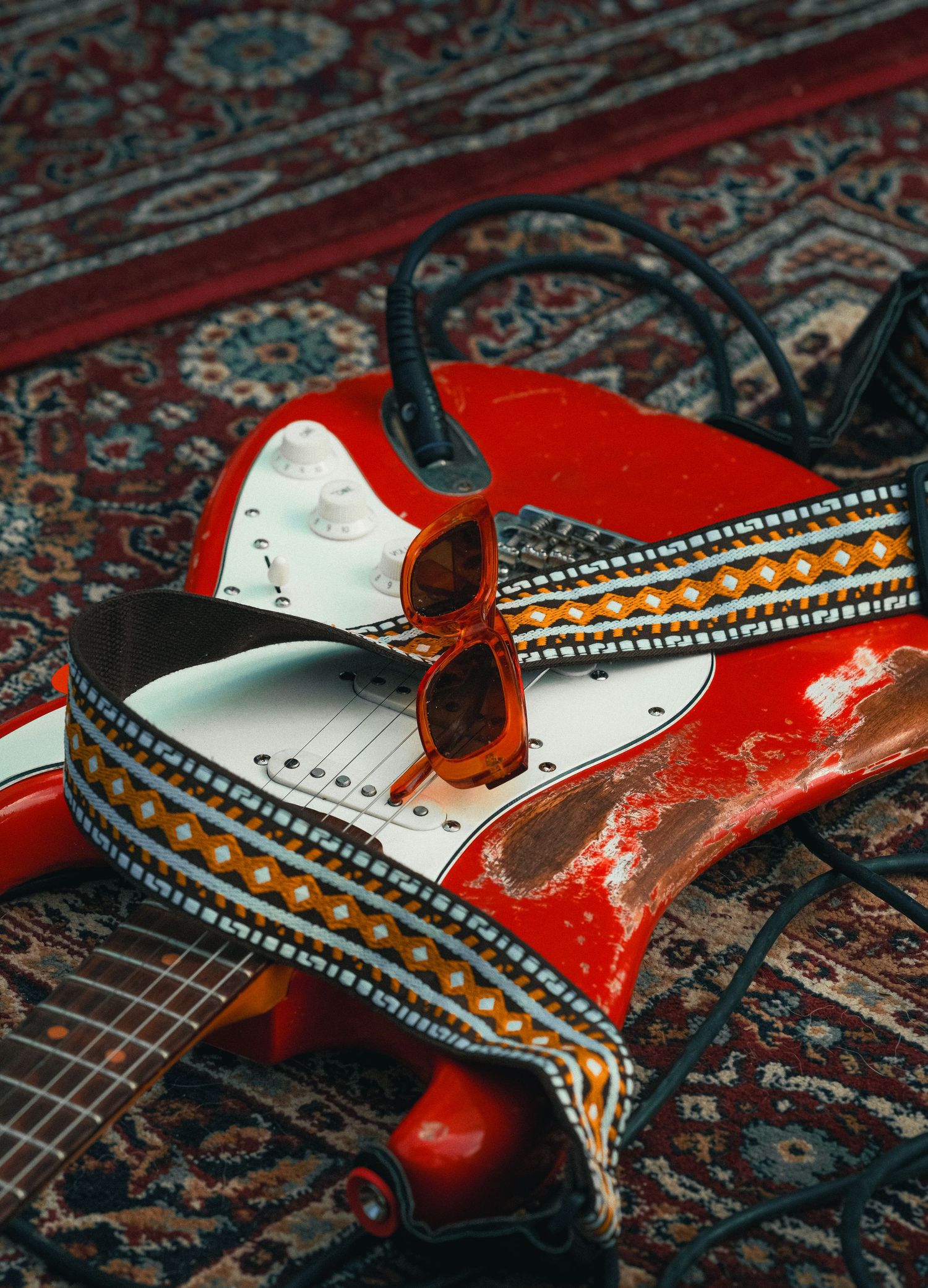 Red Fender Stratocaster electric guitar with decorative strap and sunglasses resting on a patterned rug.