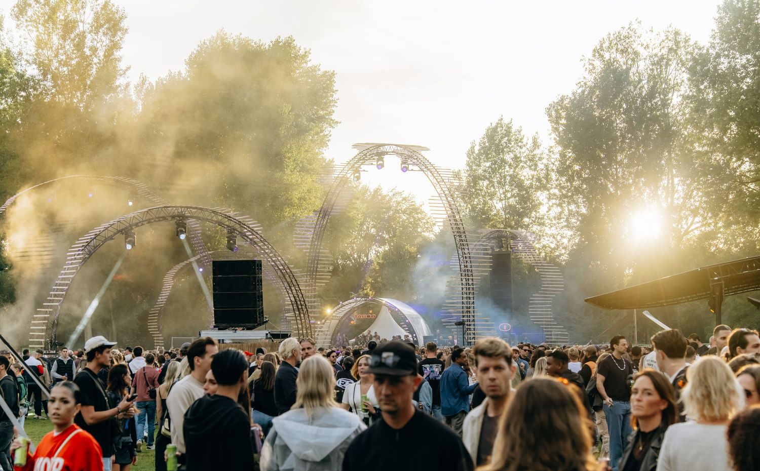 Outdoor music festival crowd with arch stage structures, smoke effects, and golden sunlight through trees.