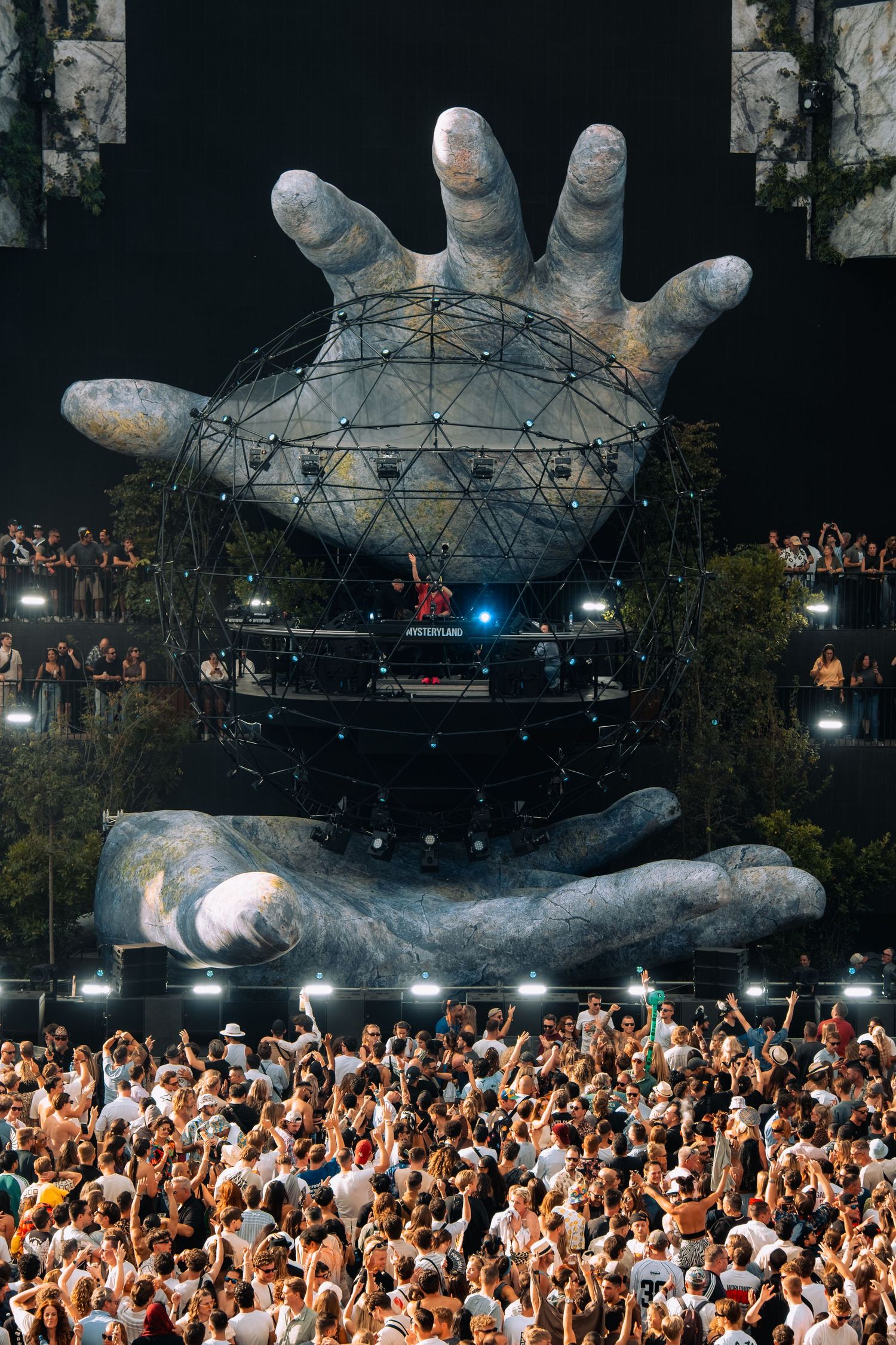 Massive crowd at Mysteryland festival facing giant sculptural hands holding a geodesic dome DJ booth stage.