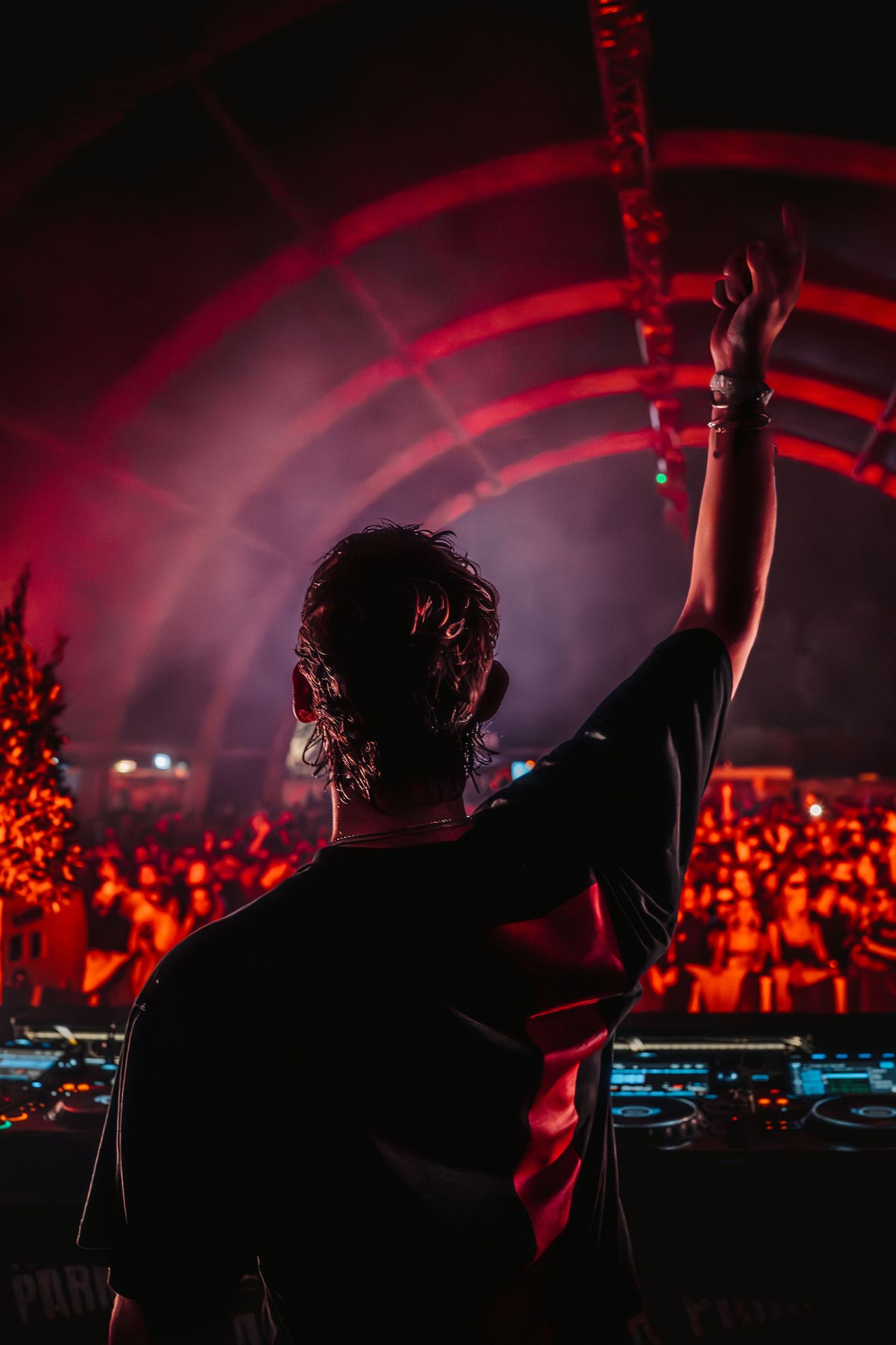 DJ with raised arm performing at a turntable setup before a large crowd in red-lit festival venue.