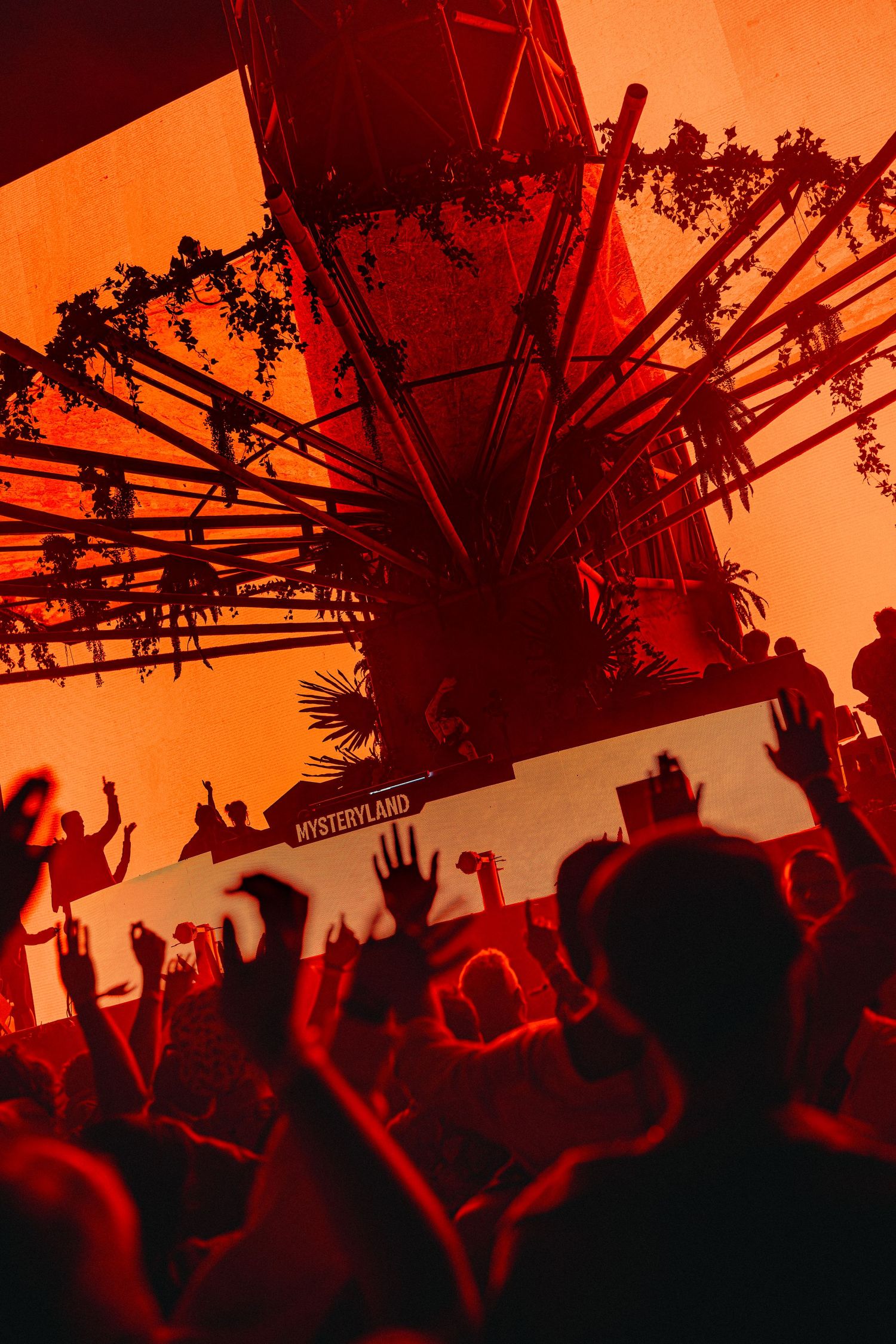 Crowd with raised hands at Mysteryland festival stage bathed in dramatic red light with silhouetted structures.