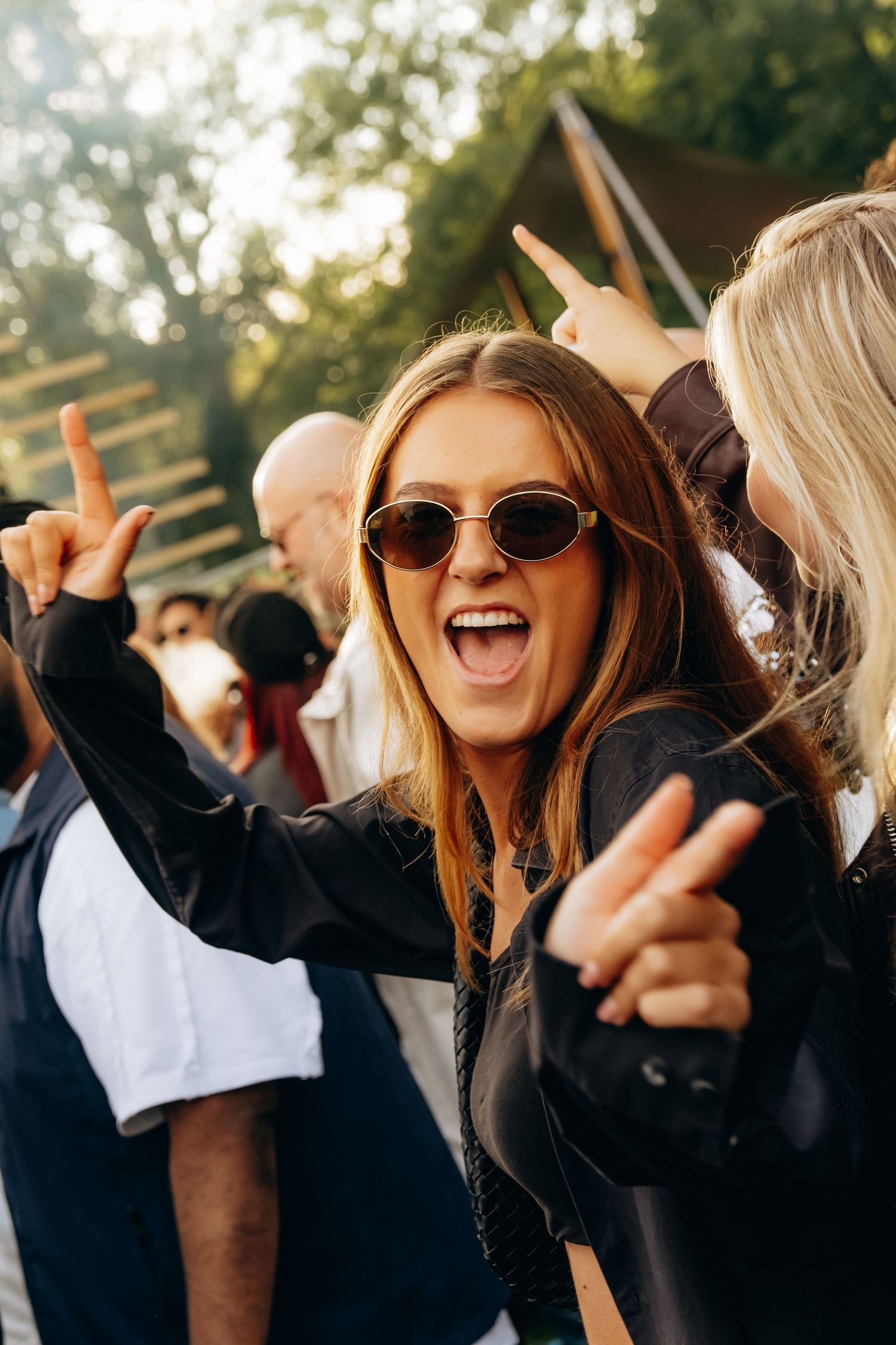 Excited young woman with sunglasses making rock signs at an outdoor music festival in golden sunlight.