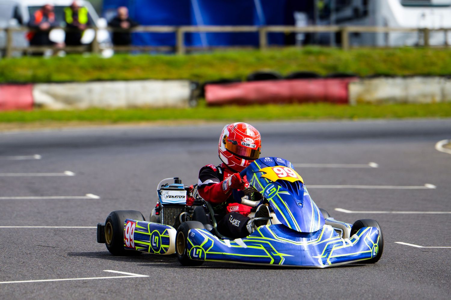 A young driver in a red helmet races a blue and yellow go-kart on an outdoor karting circuit.