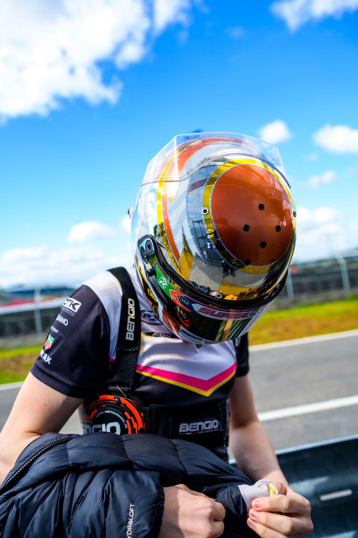 Racing driver wearing a colorful helmet and BENGIO branded racing suit at a motorsport circuit under blue skies.