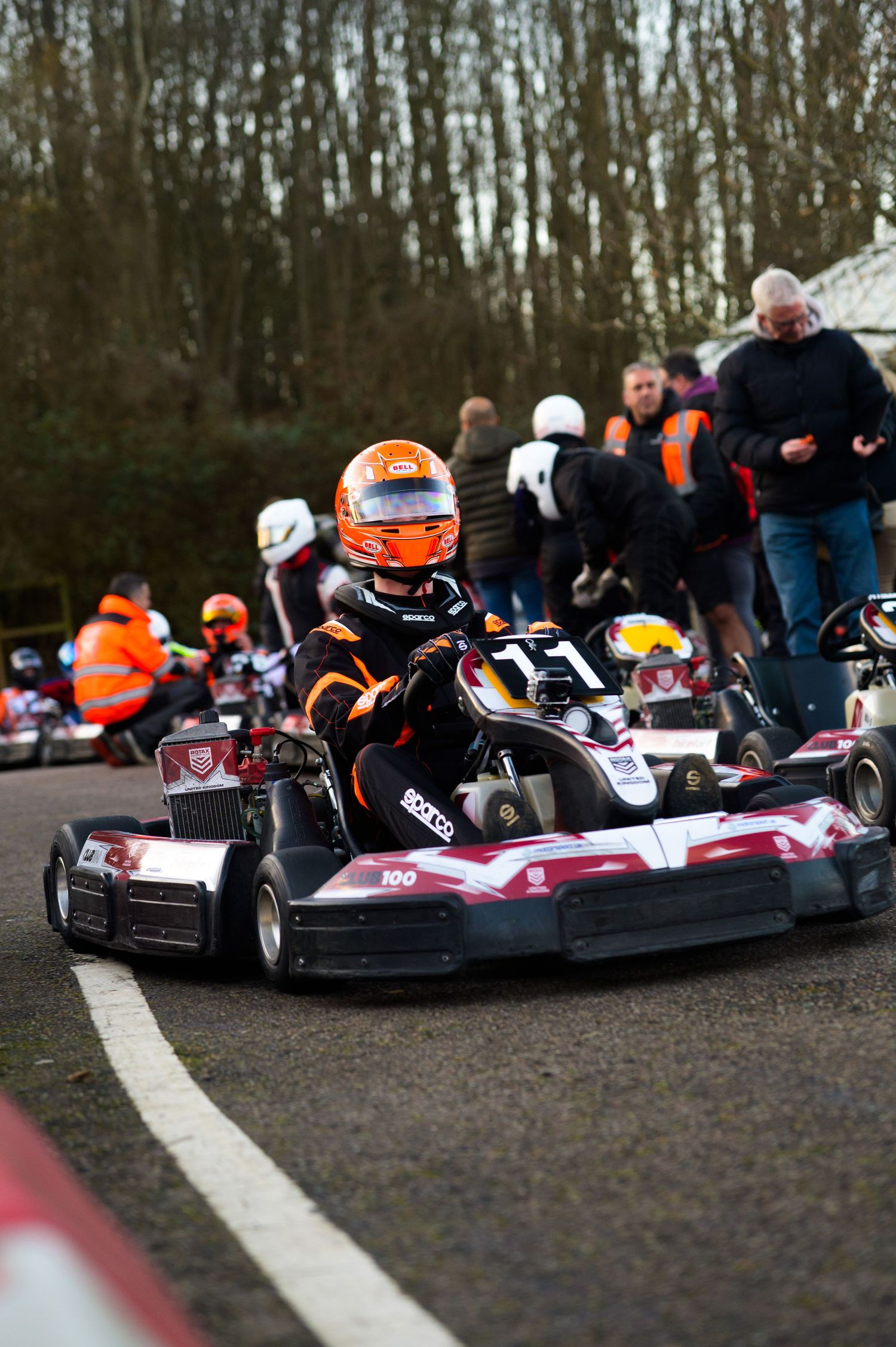 Multiple go-karts racing closely together on a karting circuit with spectators watching.