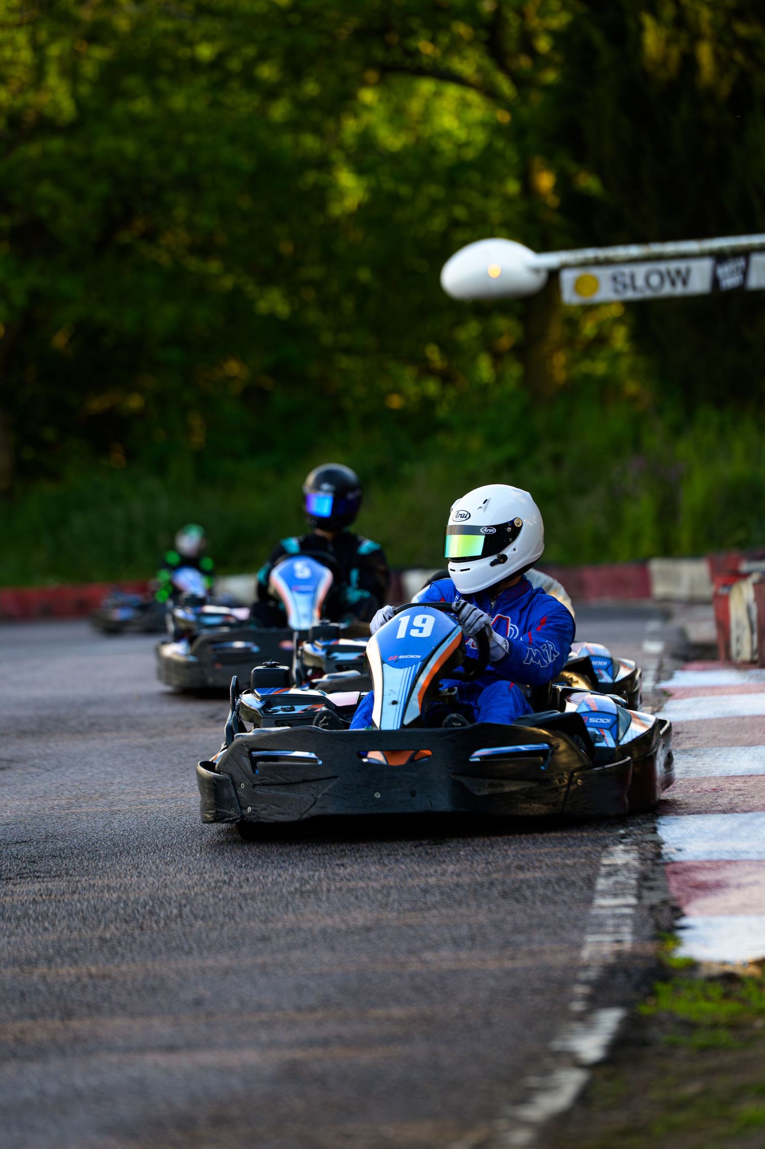 Go-kart racers in helmets compete on a wet outdoor track, with a SLOW sign visible near green trees.