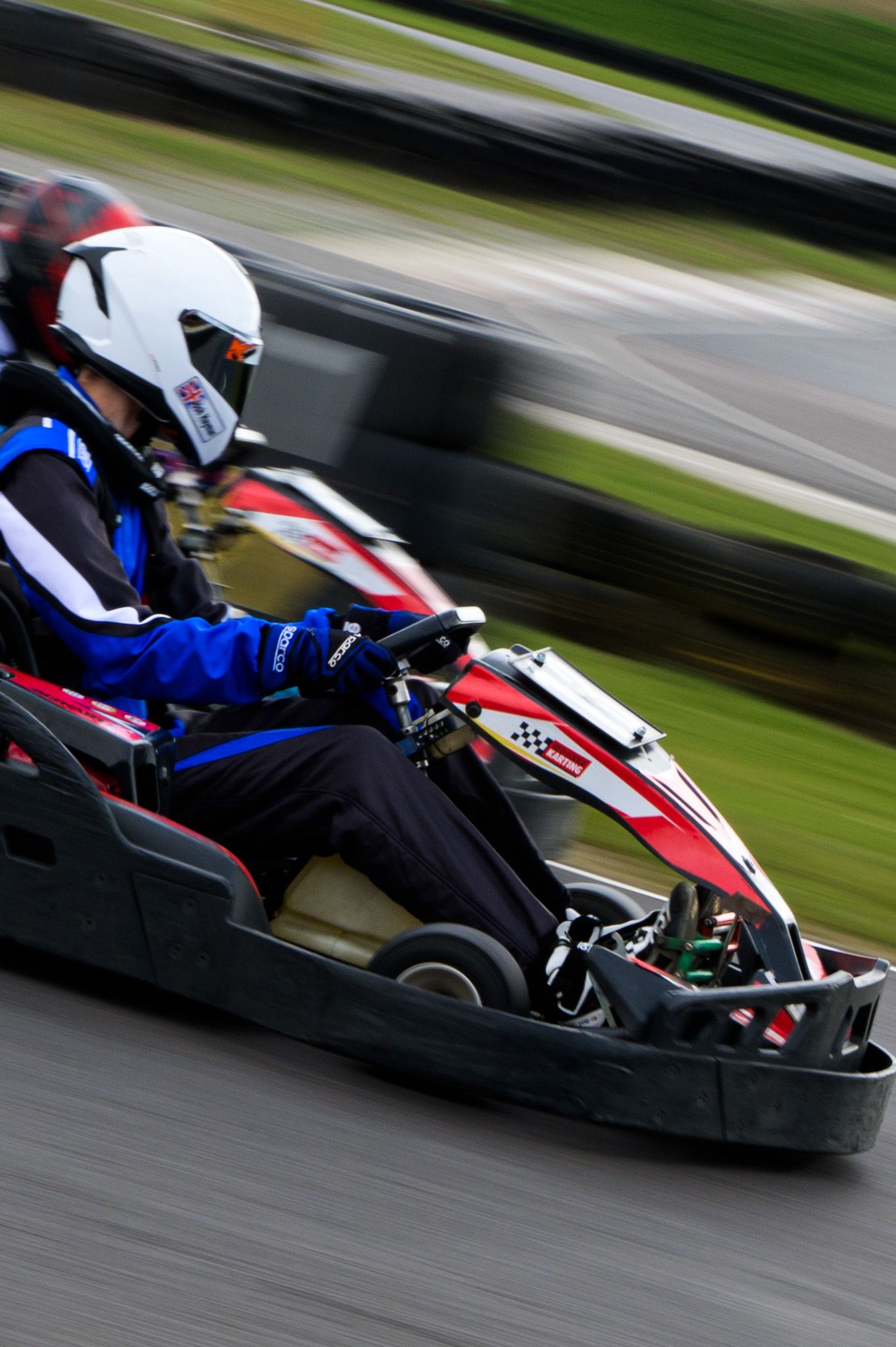 A go-kart racer in a white helmet and blue suit speeds around a racing track in a red and black kart.