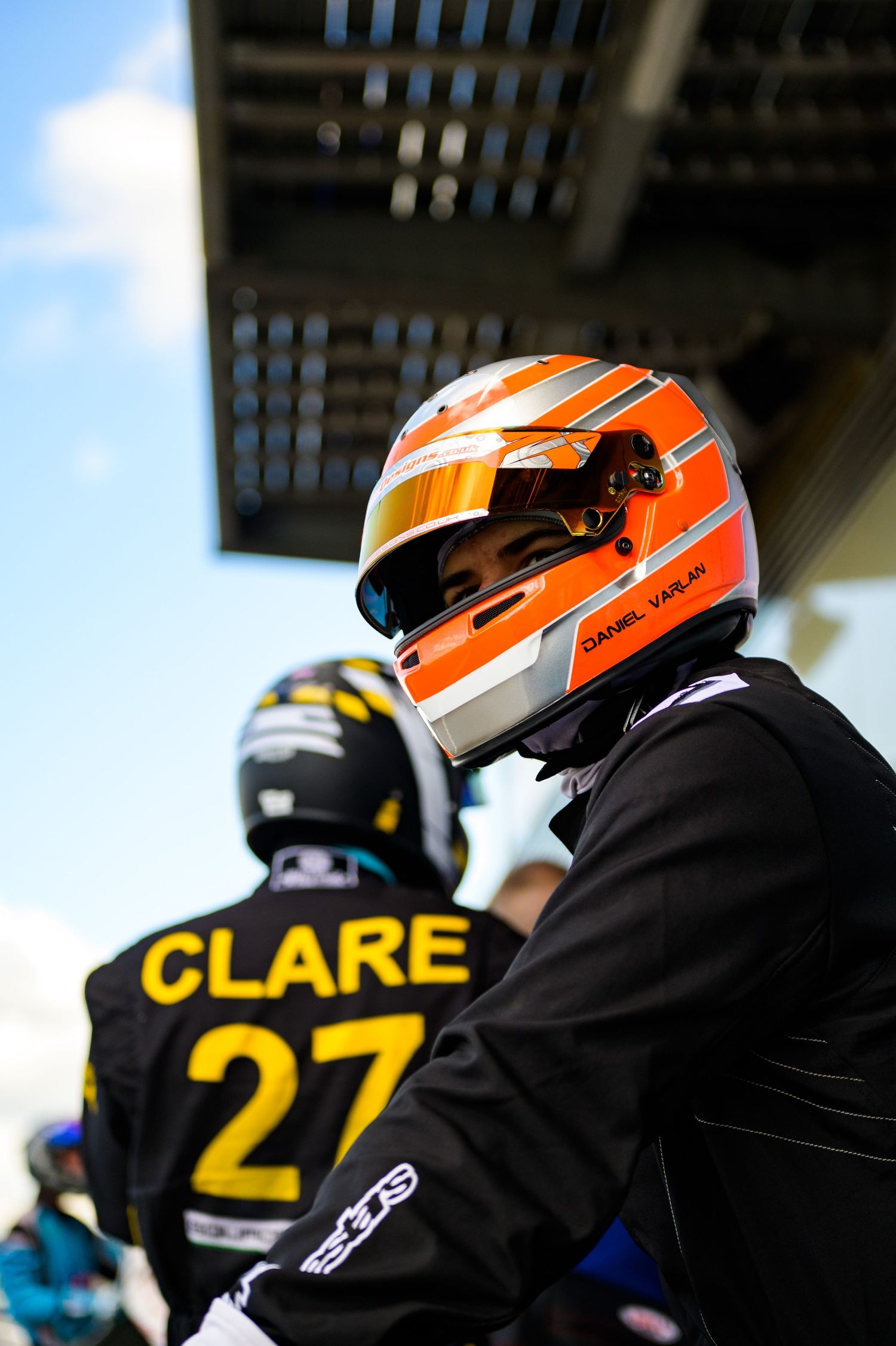 Racing driver Daniel Varlan wearing an orange and white helmet stands beside driver Clare, number 27, at a motorsport event.
