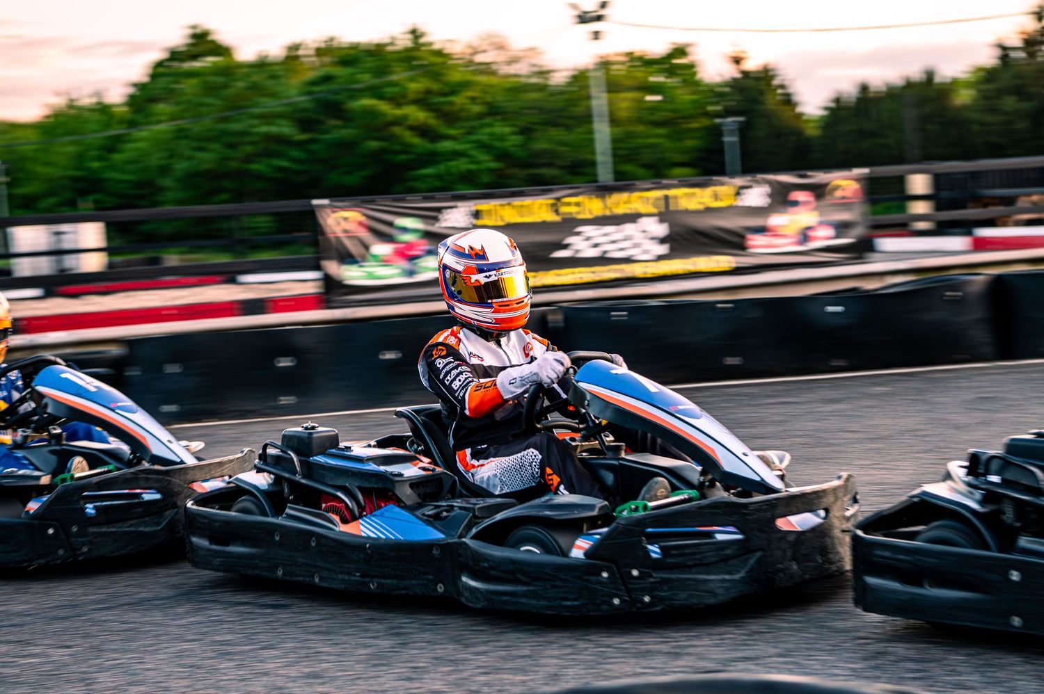 Go-kart racing on an outdoor track at dusk, with a helmeted driver leading in a blue and black kart.