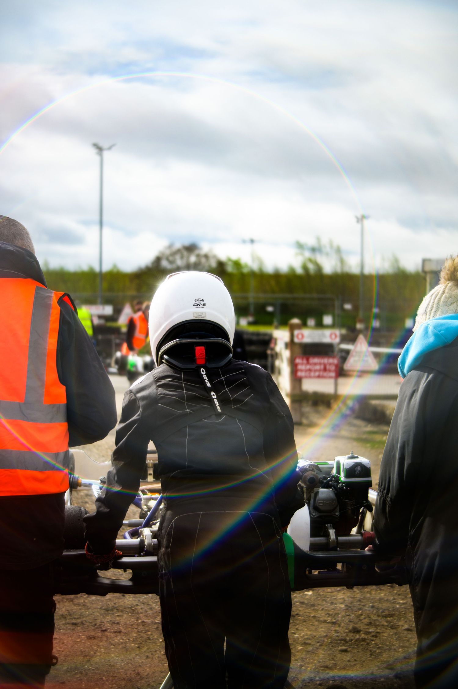A go-kart racer in a white helmet and black suit stands at a racing circuit, flanked by marshals in high-visibility vests.