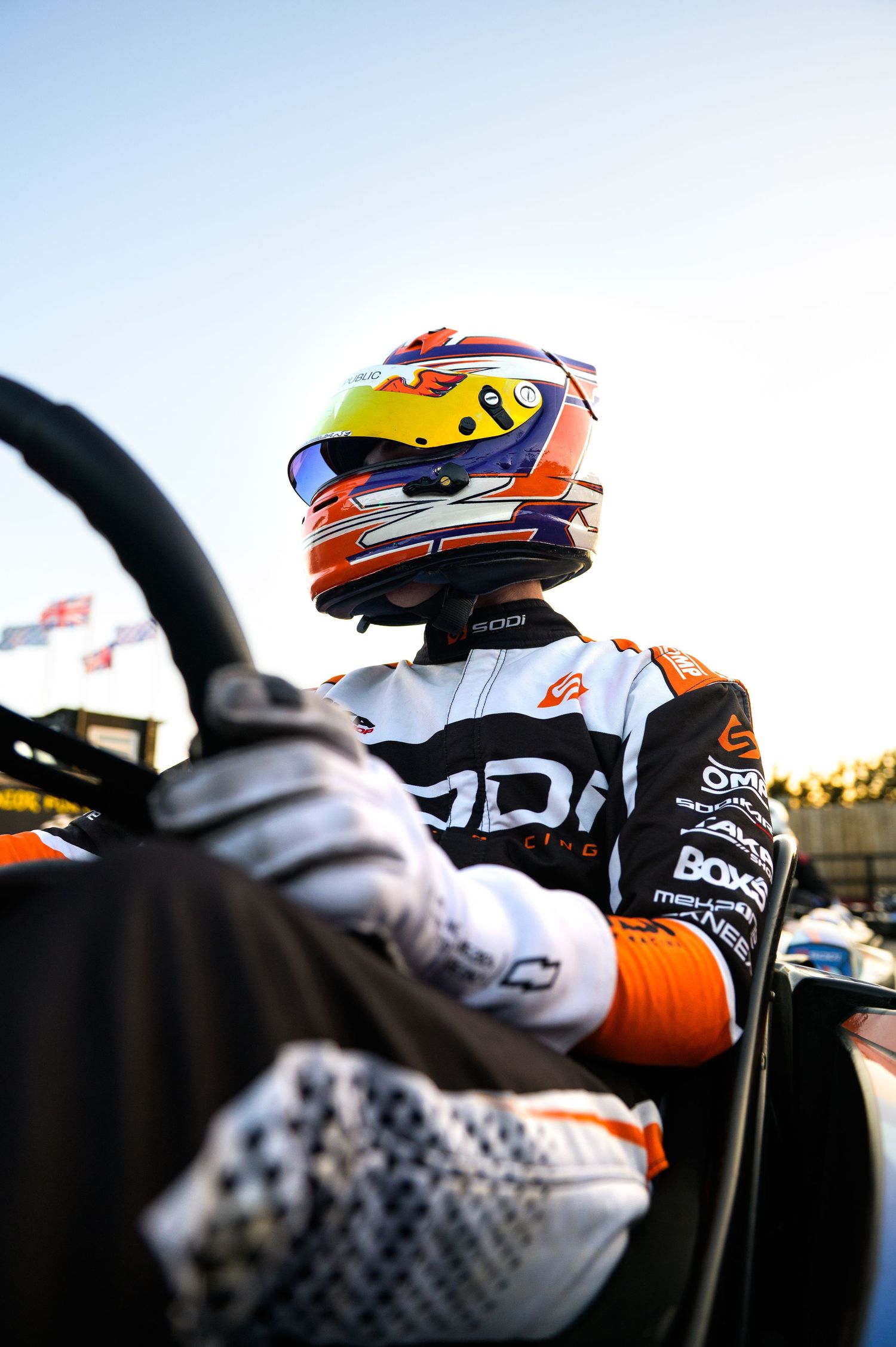 A kart racing driver in an orange and white ODD suit and colorful helmet sits ready in a racing kart.