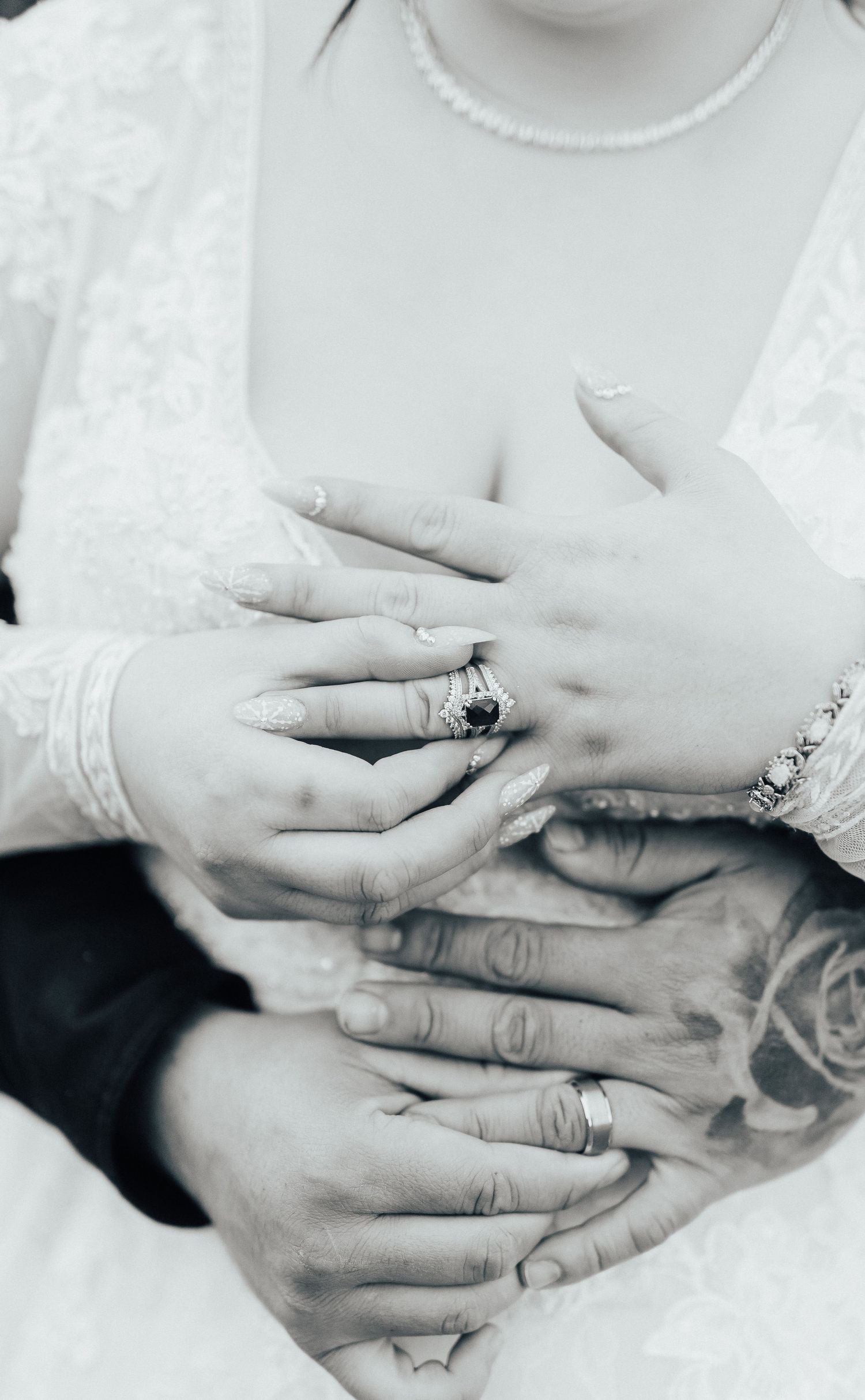 Black and white photograph of hands wearing wedding rings with delicate lace sleeves and a pearl necklace.