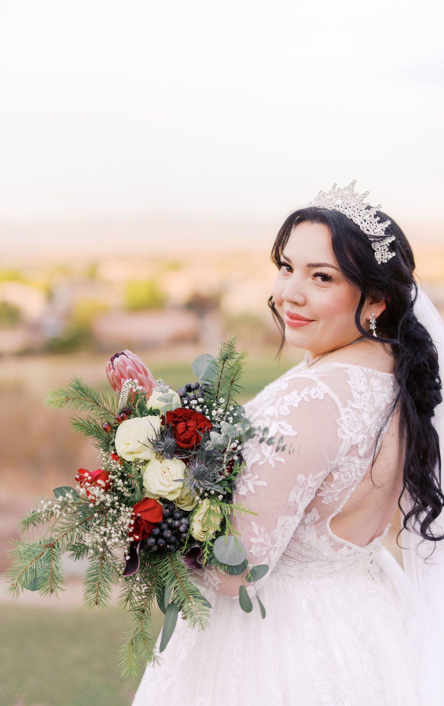 A bride in a lace wedding dress holds a stunning winter bouquet with red and white flowers, pine sprigs and berries.