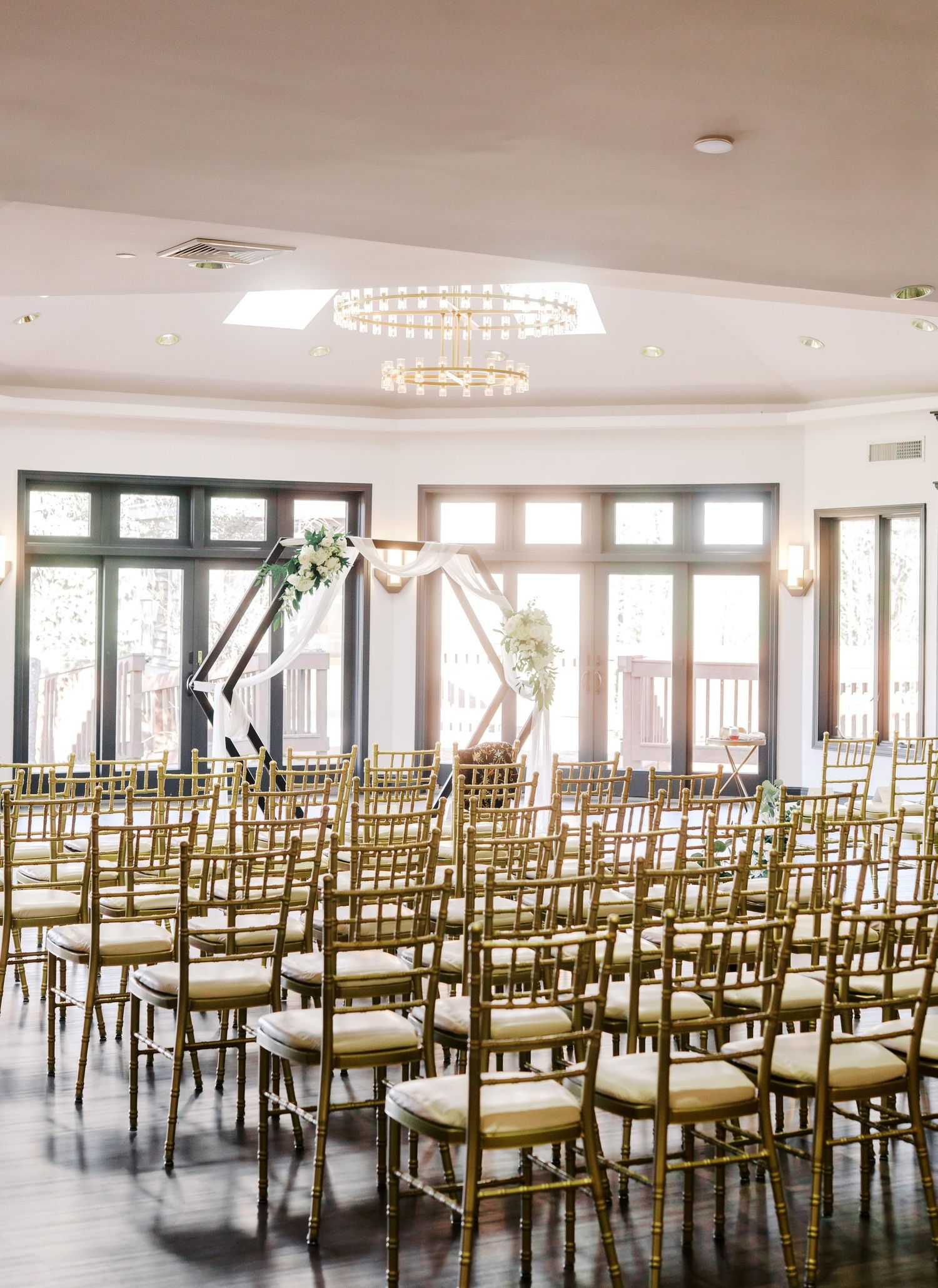 Elegant wedding ceremony setup with rows of gold chairs and large windows in a bright reception hall.