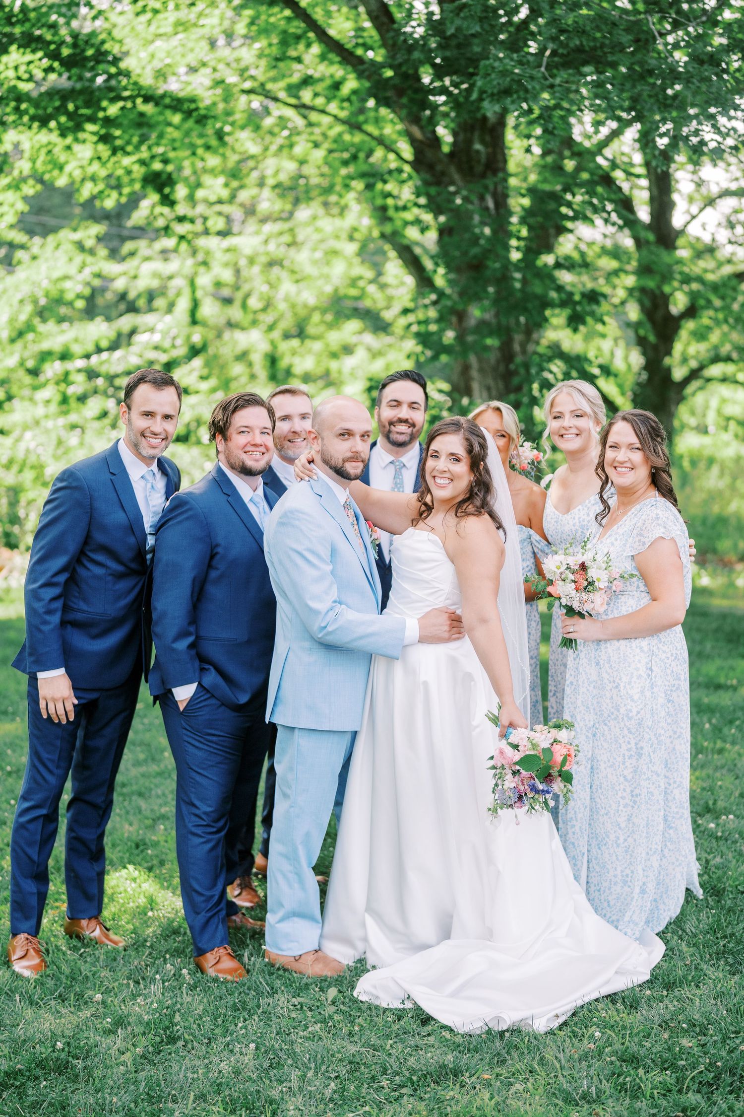 Wedding party posing casually on green lawn in coordinated blue and white outfits.