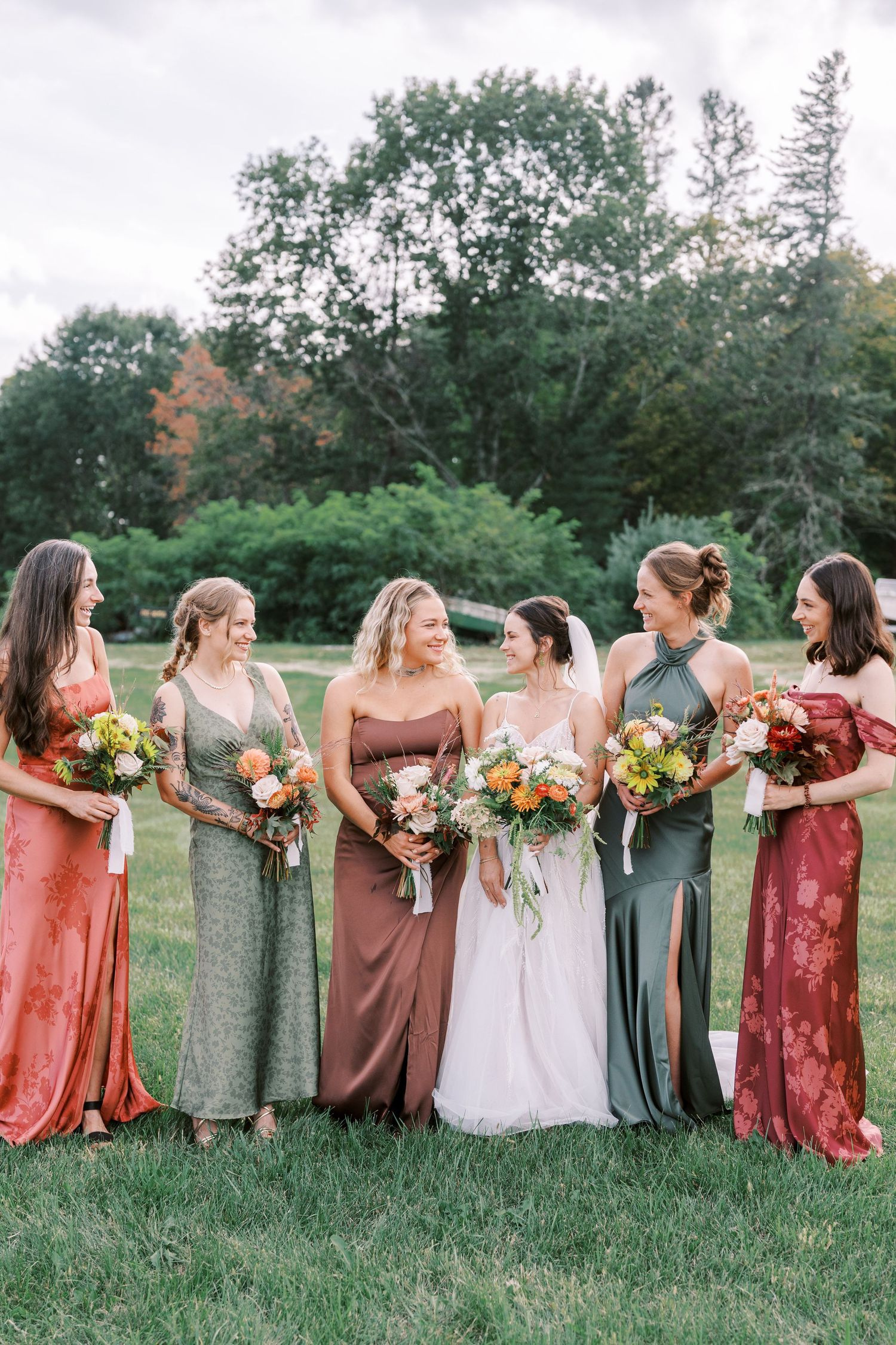 Bridal party in mismatched dresses of rust, green and burgundy colors poses together on grassy field.