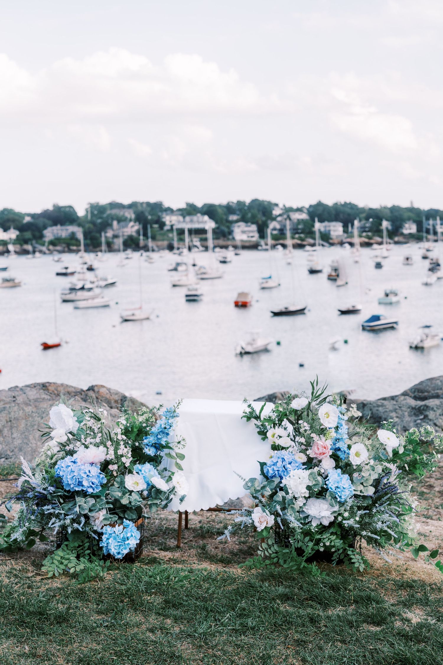 Coastal view with boats in harbor and elegant blue and white floral arrangements along rocky shoreline.