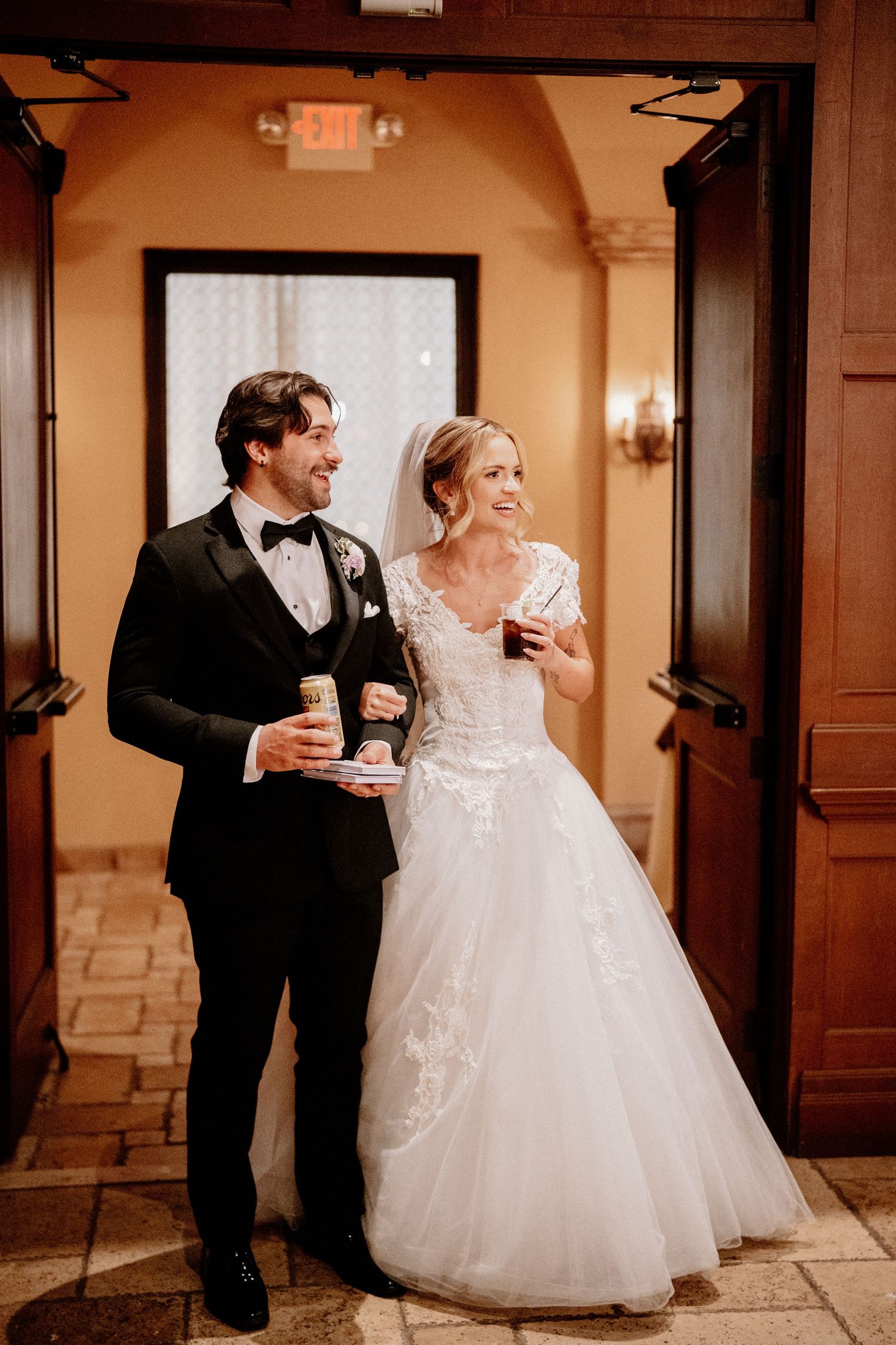 Newlywed couple shares a playful moment in an elegant hallway while holding beverages at their wedding reception.
