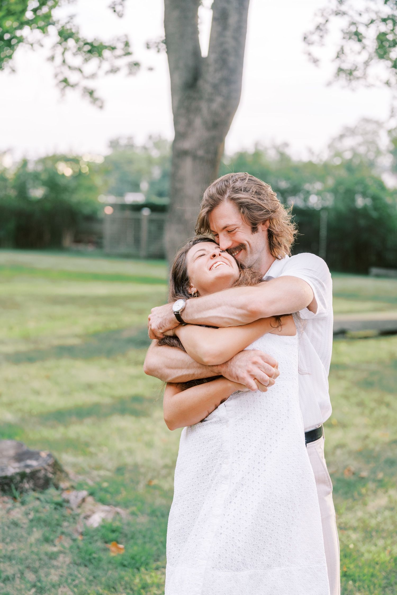 A couple embraces in a sunny garden setting with trees in the background.