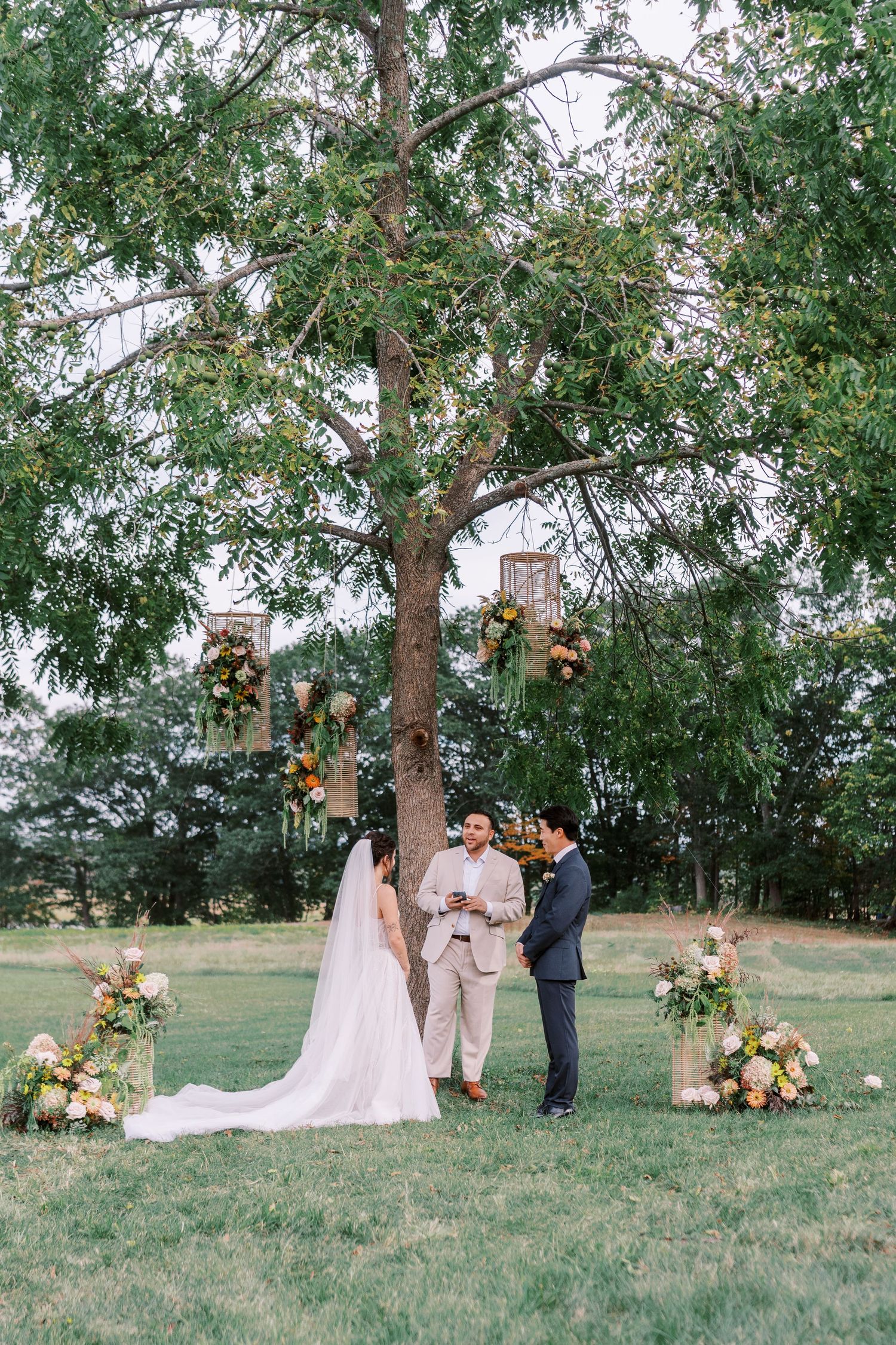 Outdoor wedding ceremony under tall trees with hanging lanterns and floral arrangements.