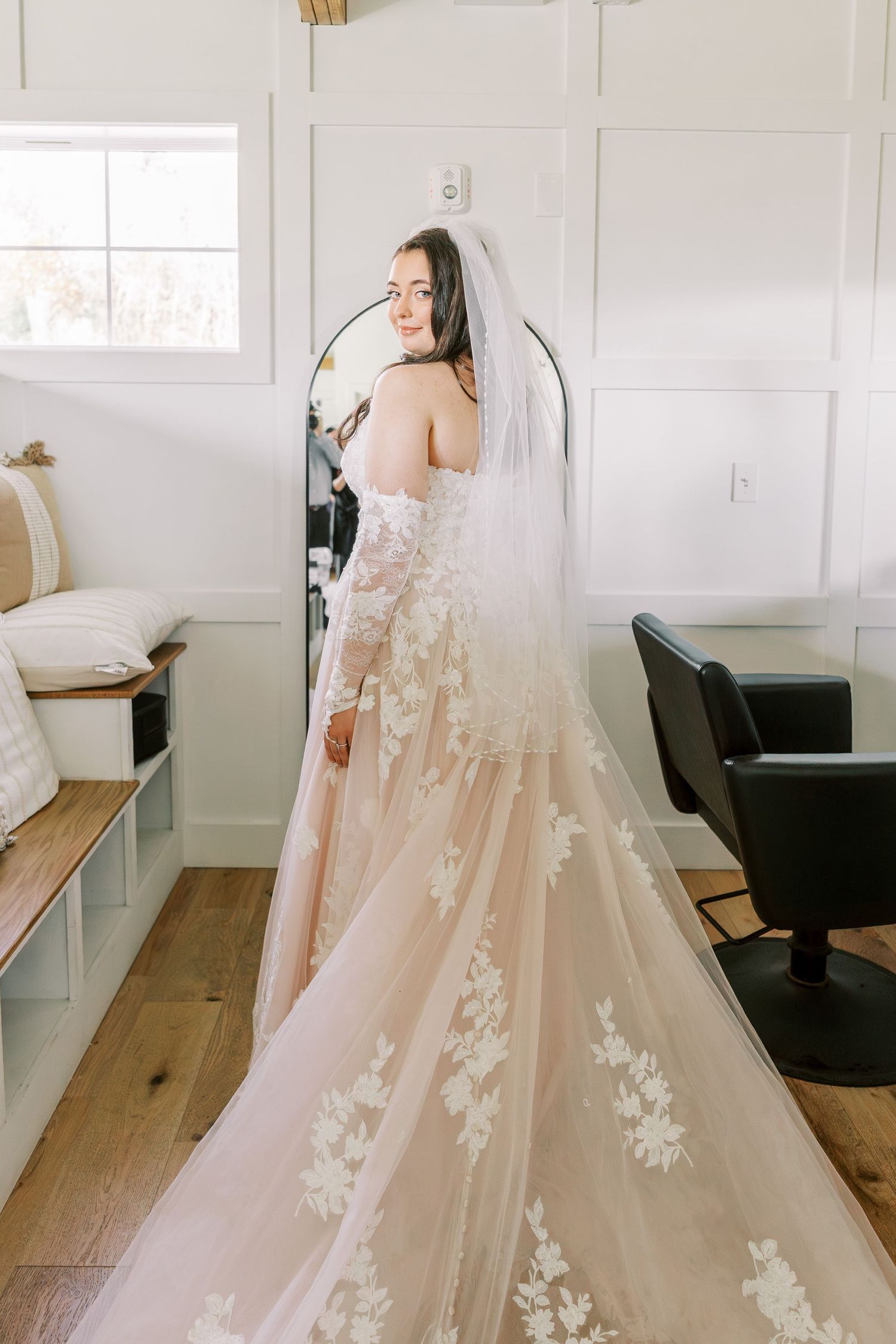 A bride in an elegant off-shoulder wedding gown with floral lace appliques and flowing veil stands in a bright white room.