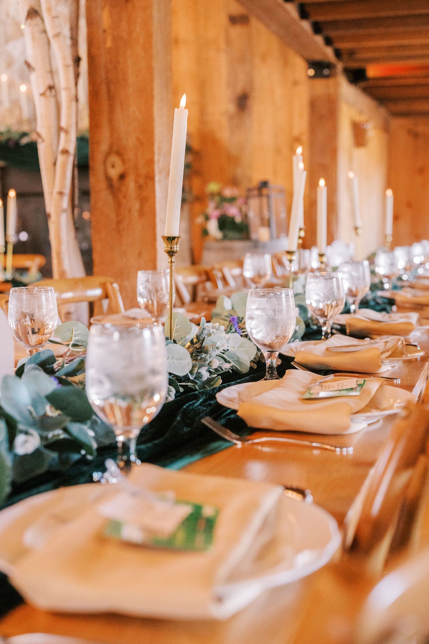 Elegant table setting at rustic barn wedding reception with white candles and greenery garland along wooden tables.