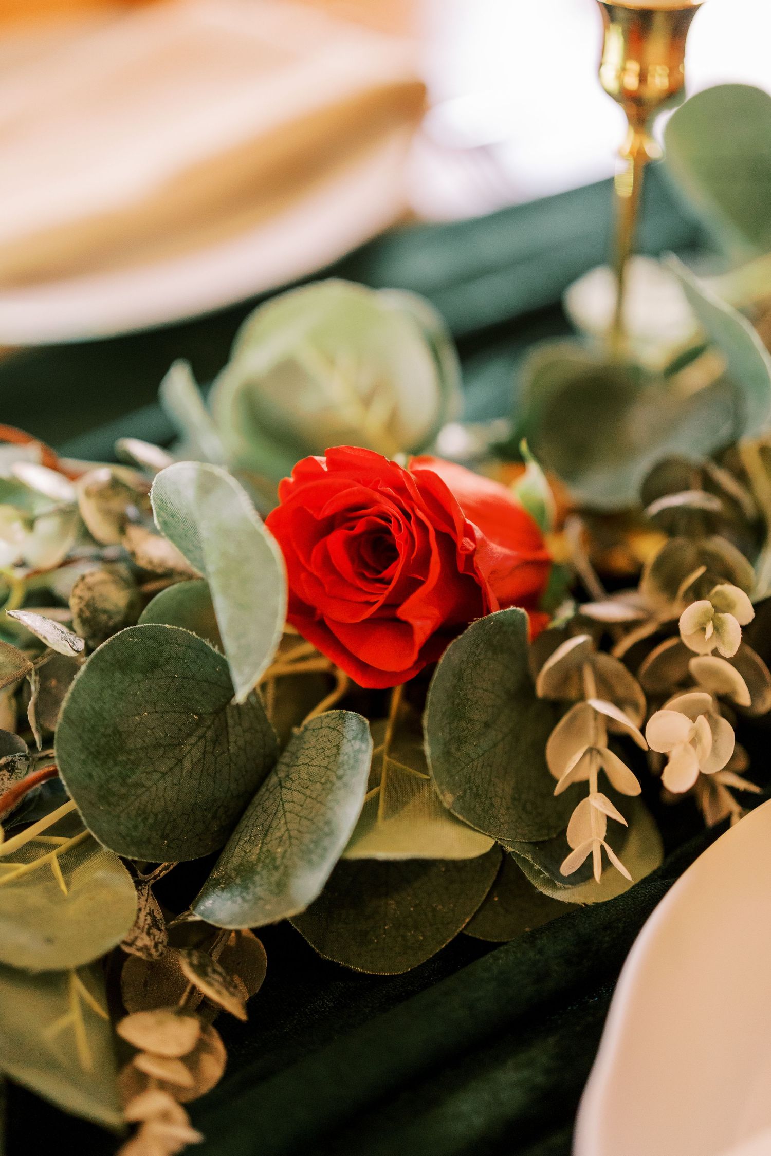 Red roses and eucalyptus leaves create an elegant table centerpiece with gold candlesticks for wedding decor.