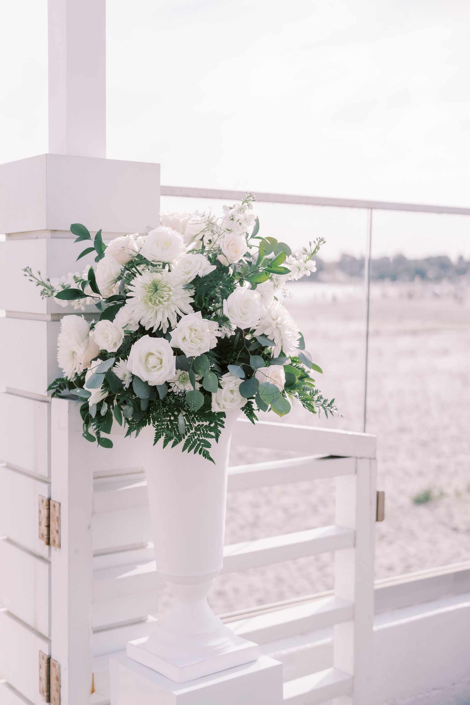 White floral arrangements with roses and greenery decorate a white railing overlooking a scenic outdoor view.