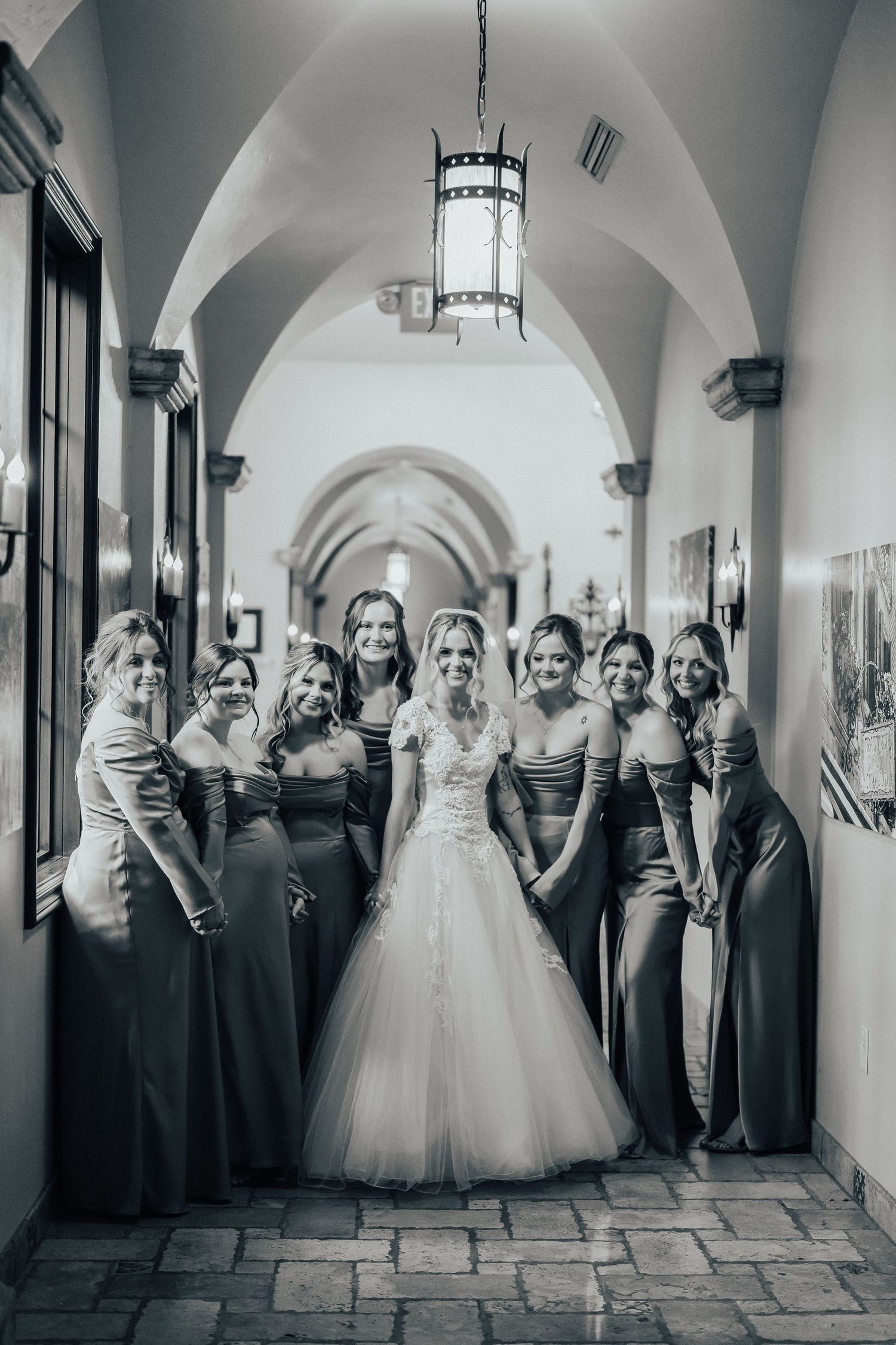 A black and white wedding party photo in an elegant arched hallway with stone floors and hanging lanterns.