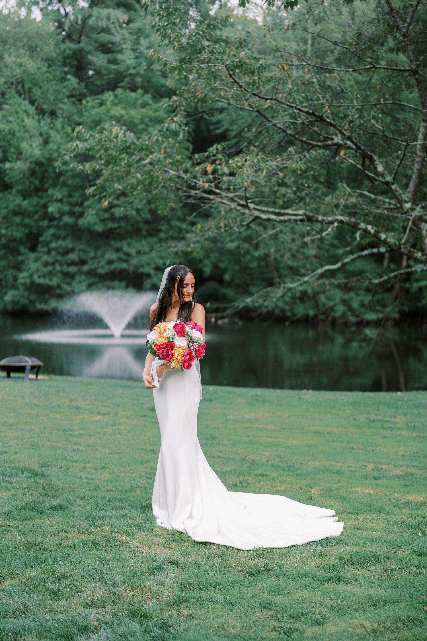 Bride in flowing white dress holds colorful bouquet by lakeside with fountain.