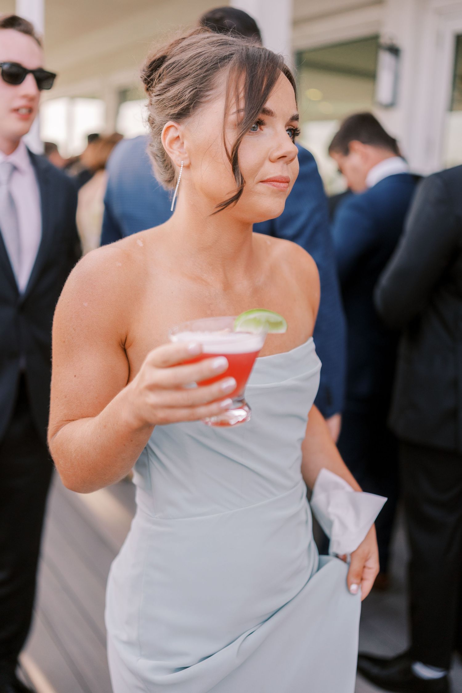A woman in a strapless gray dress holds a red cocktail with lime garnish at an elegant event.
