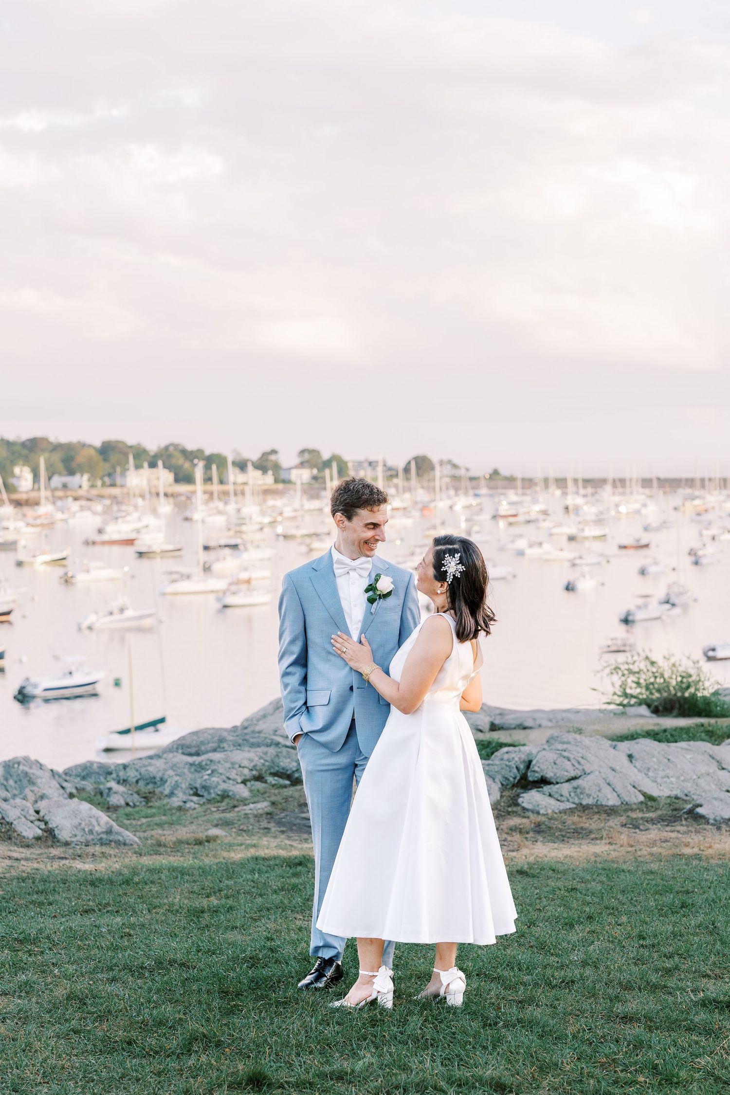 Romantic wedding couple sits along grassy waterfront near scenic marina filled with boats during sunset.