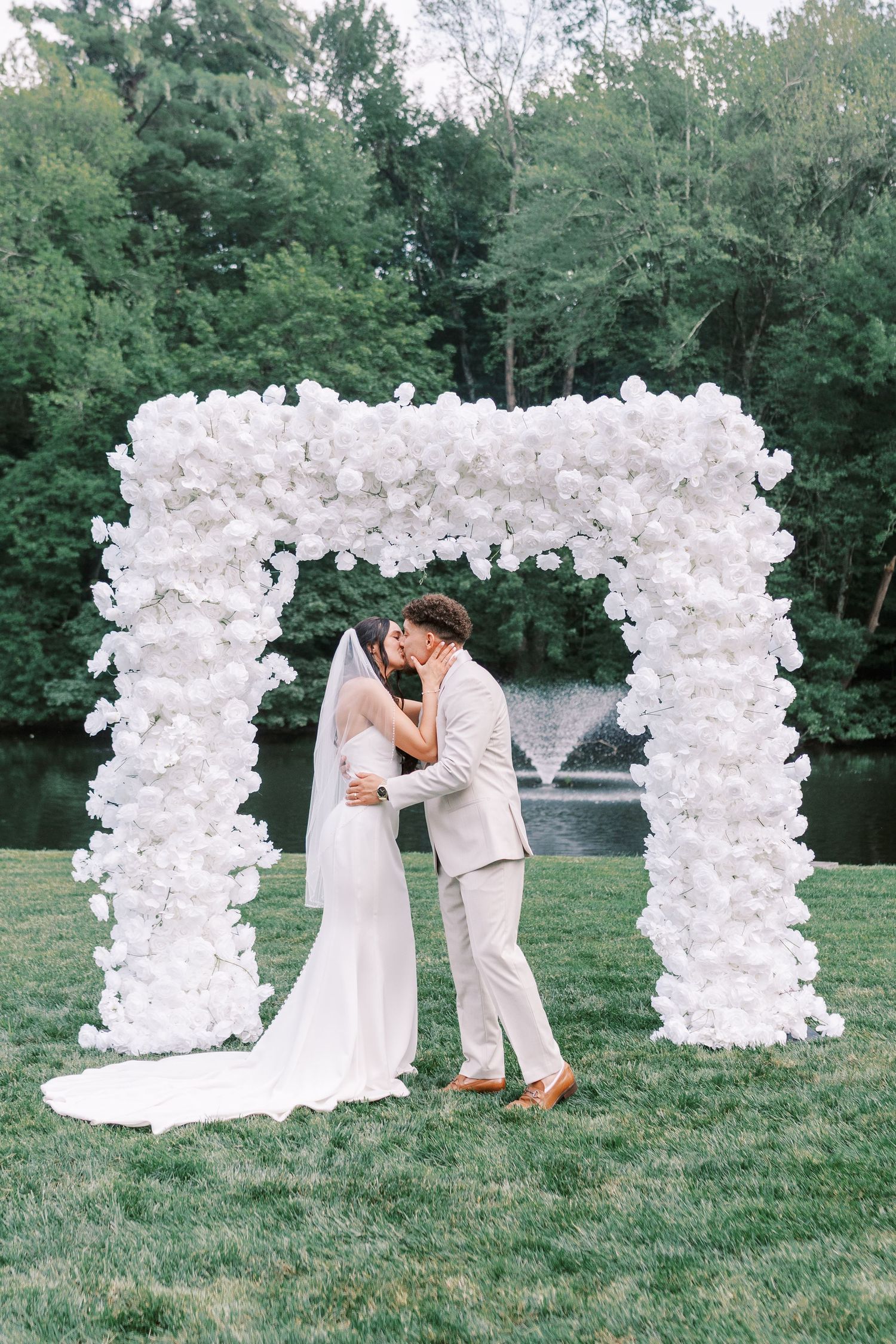 Newlyweds share a romantic moment under an elaborate white floral arch at their outdoor wedding.