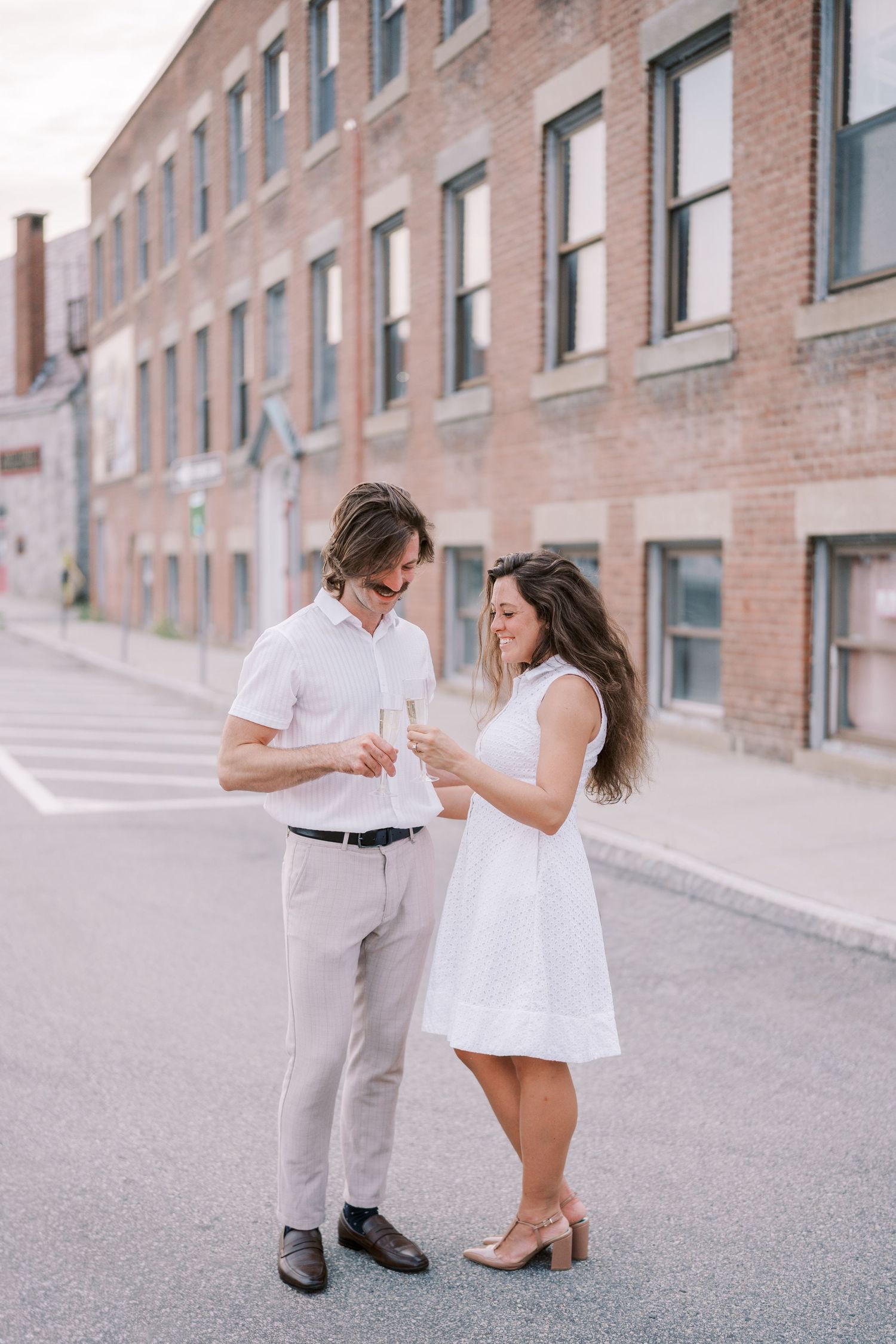 A couple in white and beige outfits share a moment on a brick-lined street.