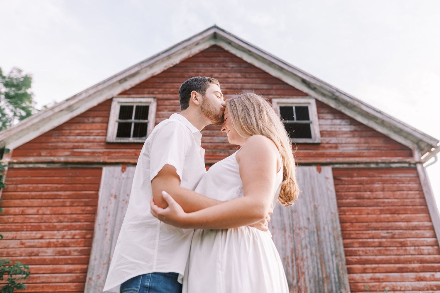 A couple embraces in front of a rustic red barn on a summer day.