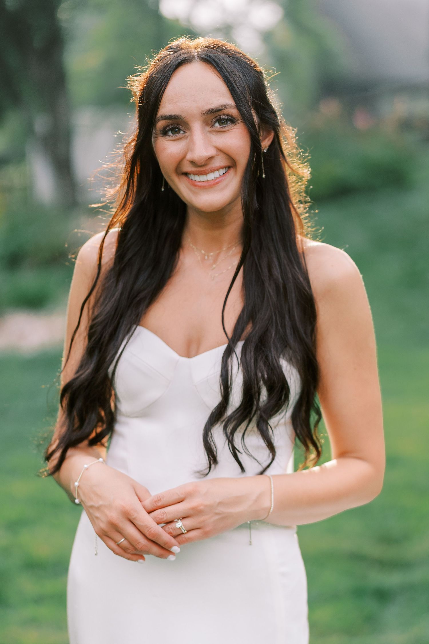 A bride in a strapless white wedding dress smiles outdoors with long dark hair and natural backlit glow.