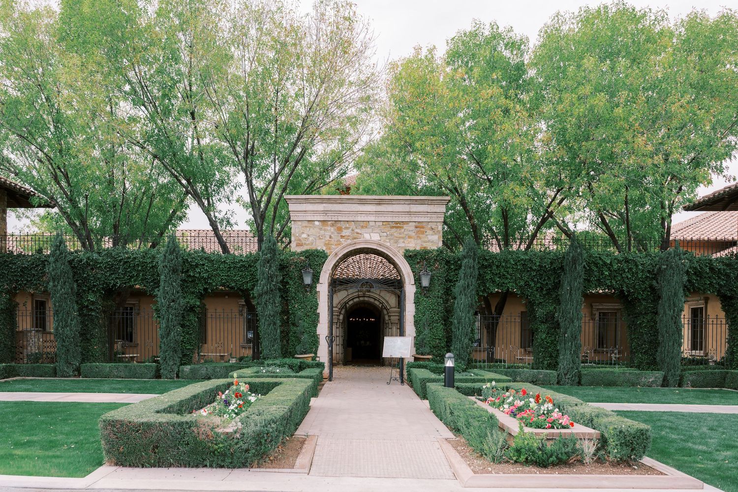 Elegant courtyard entrance with arched doorway, manicured hedges, and green lawn at a brick building with tall trees.