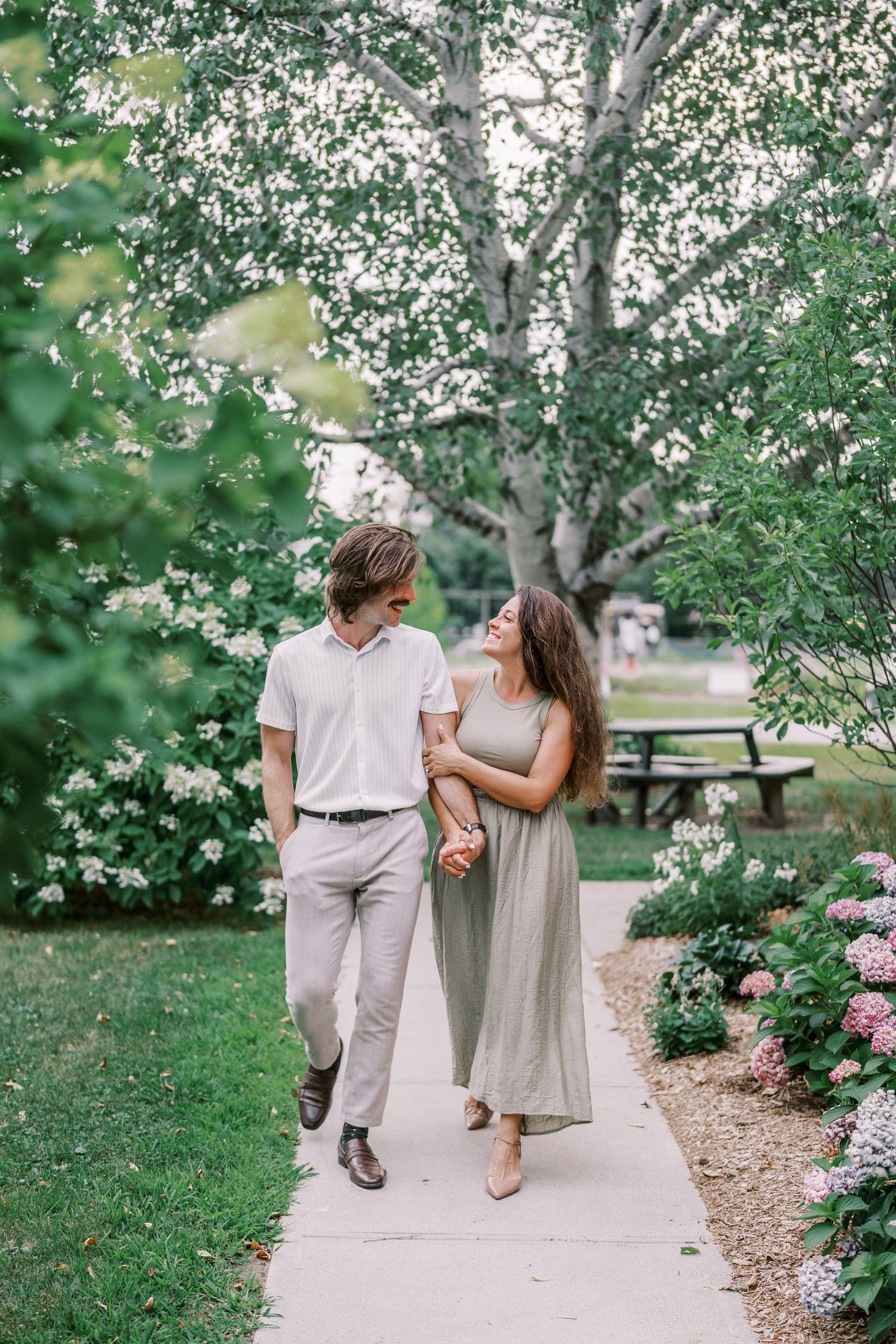 A couple walks hand in hand along a garden path surrounded by blooming trees and pink flowers.