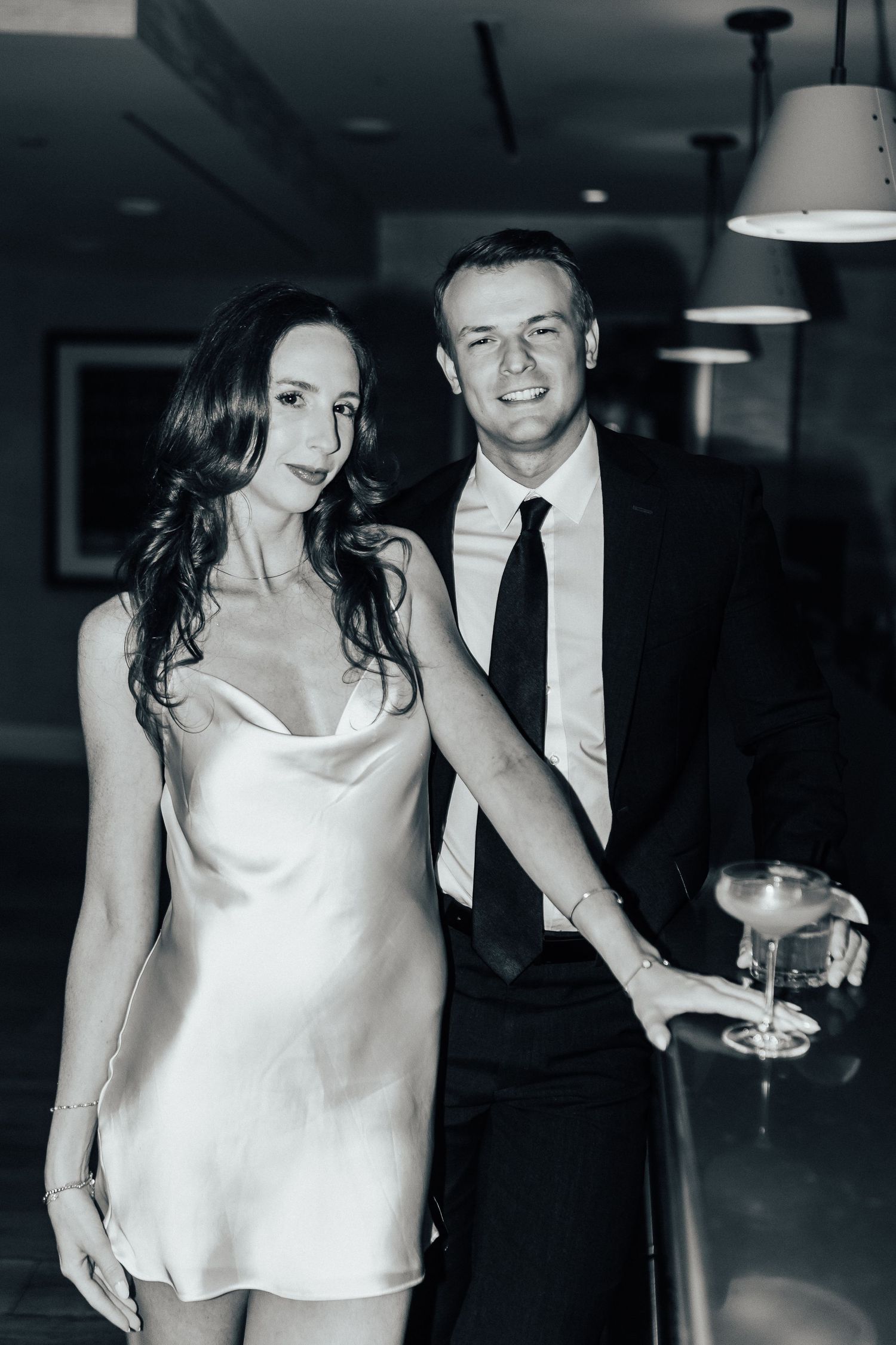 A couple celebrates at a formal event, with the woman wearing a white silk slip dress and the man in dark attire.