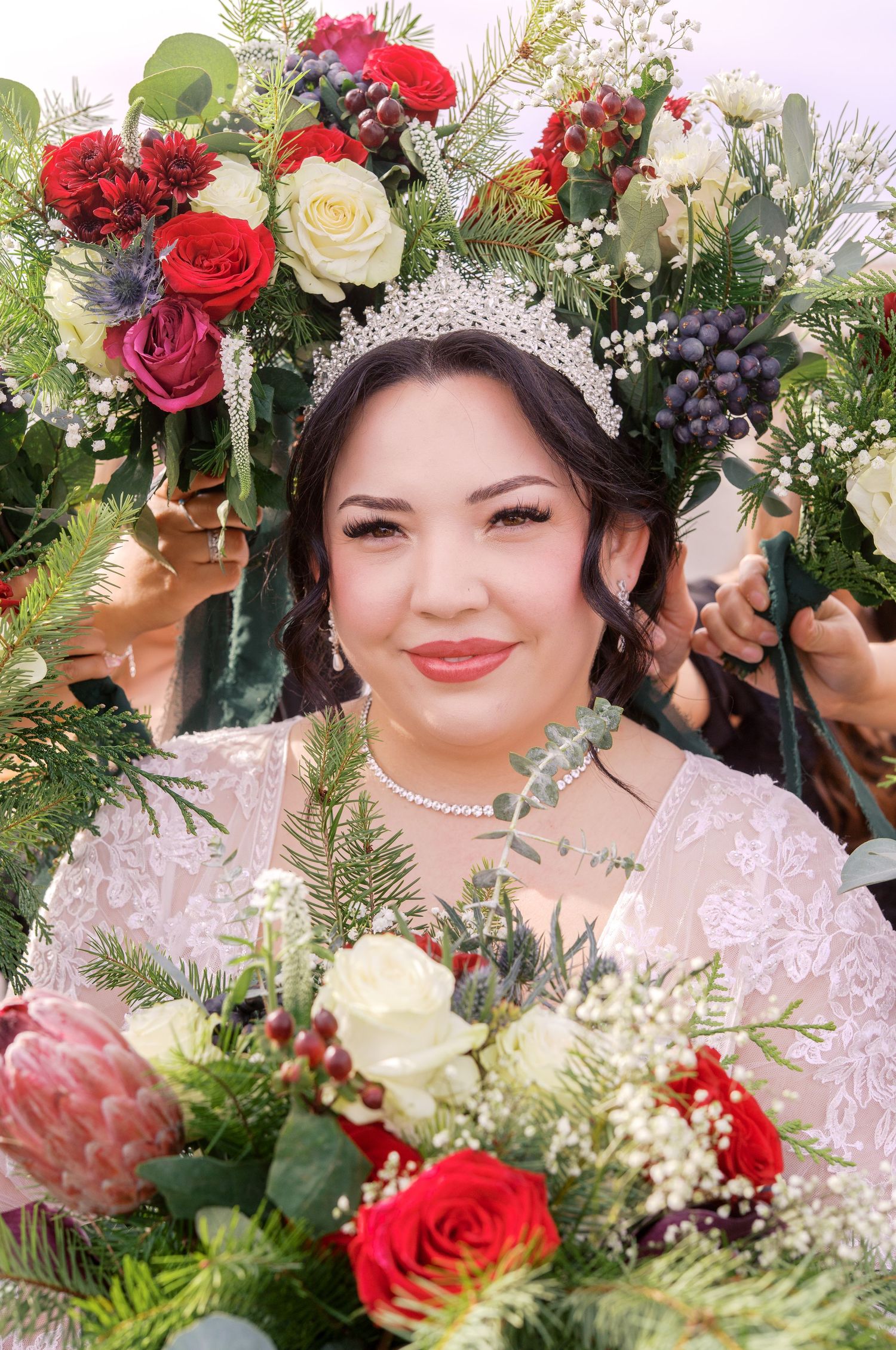 Beautiful bridal portrait surrounded by vibrant red roses, white flowers and greenery with delicate pearl crown and lace details.