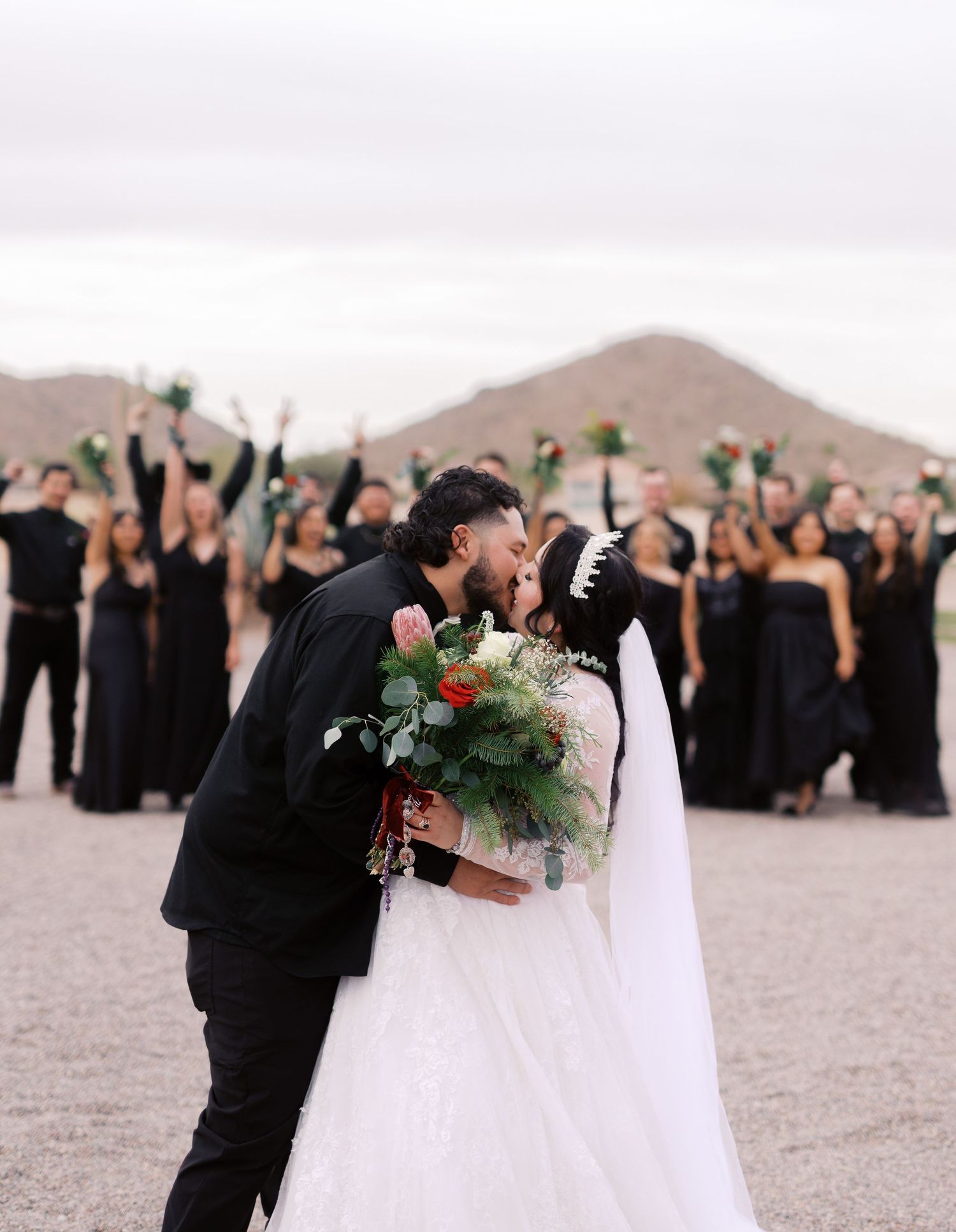 Wedding couple share a kiss in the desert with bridesmaids in black dresses holding greenery in the background.