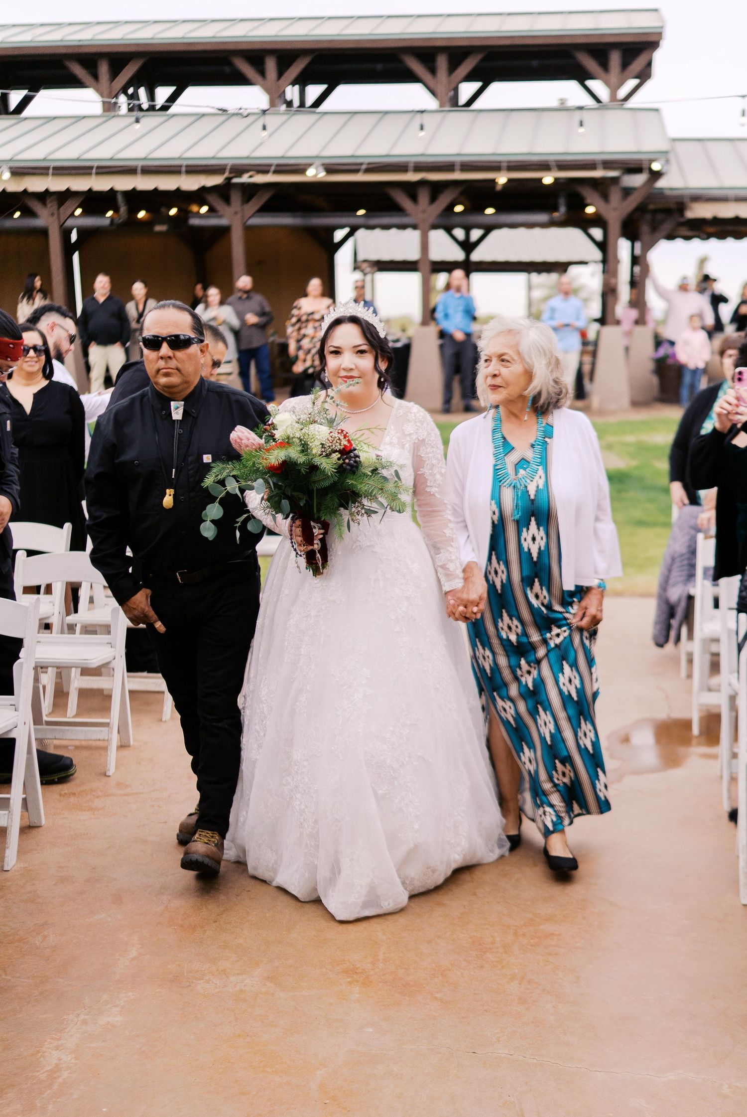 A wedding ceremony procession takes place under a covered outdoor pavilion with guests seated on white chairs.