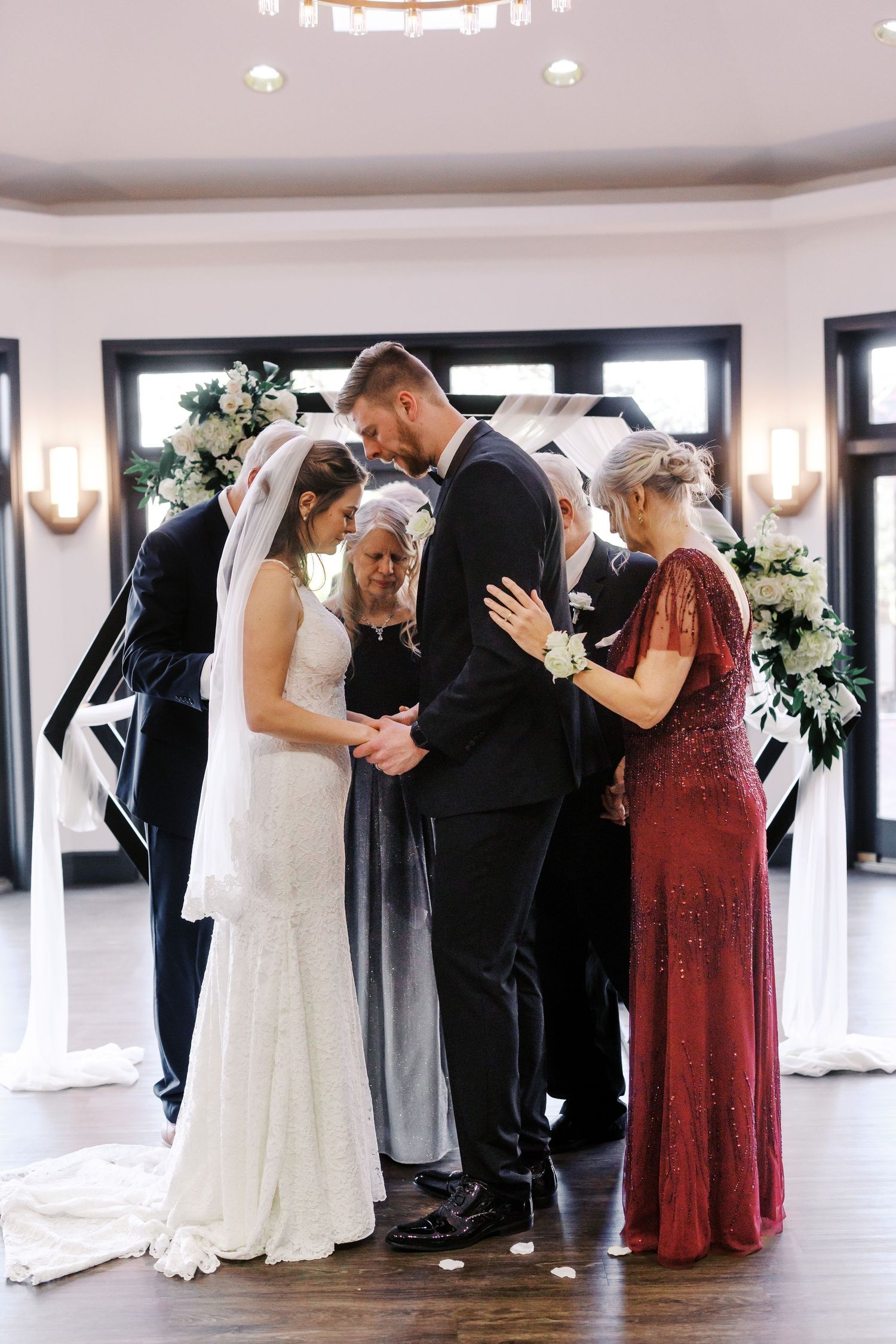 Wedding ceremony with a couple exchanging vows surrounded by family in a bright venue with floral decor.