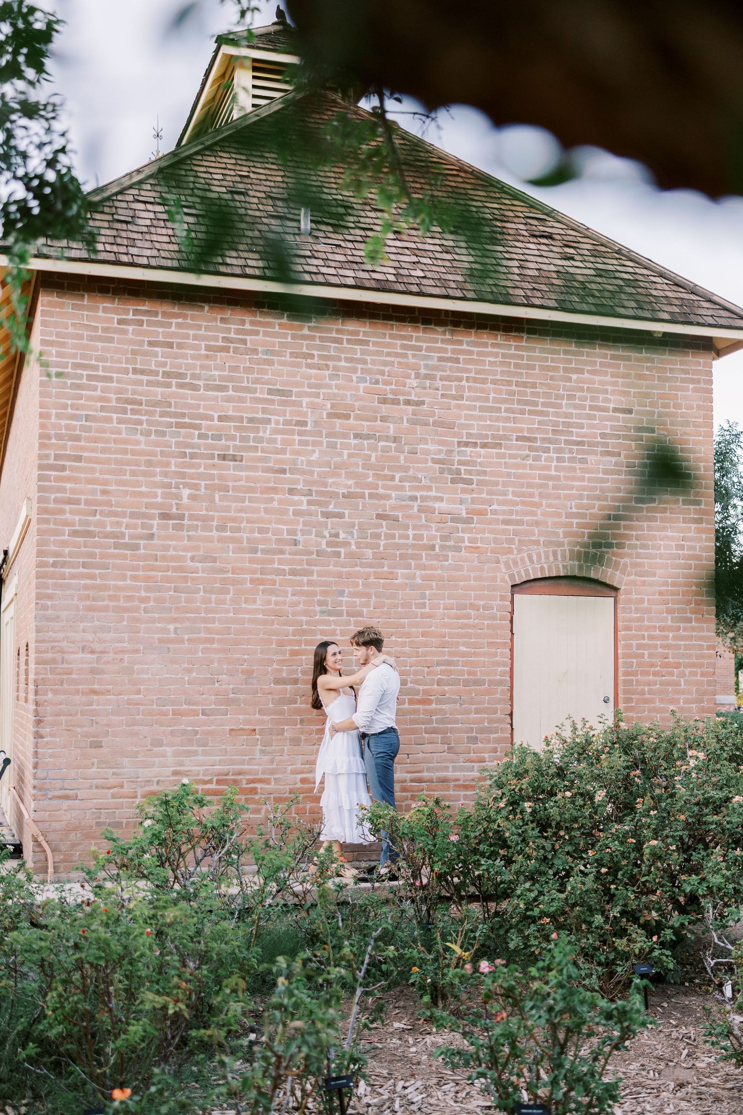 A couple walking together near a pink brick building with green shrubs in the foreground.