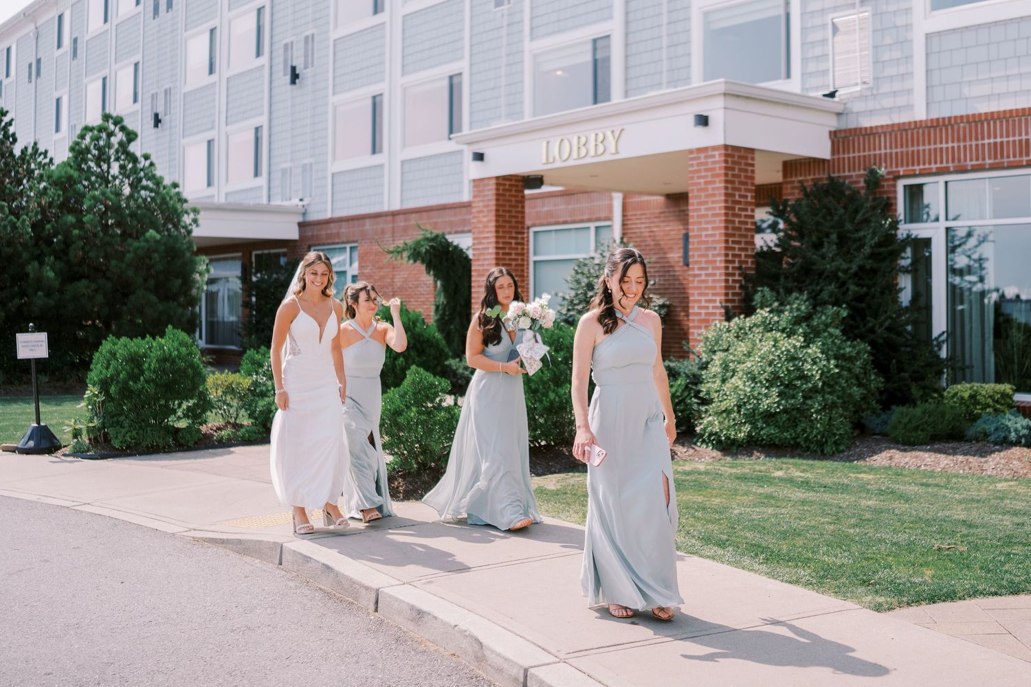 A wedding party walks together outside a brick building on a sunny day wearing light gray dresses and suits.