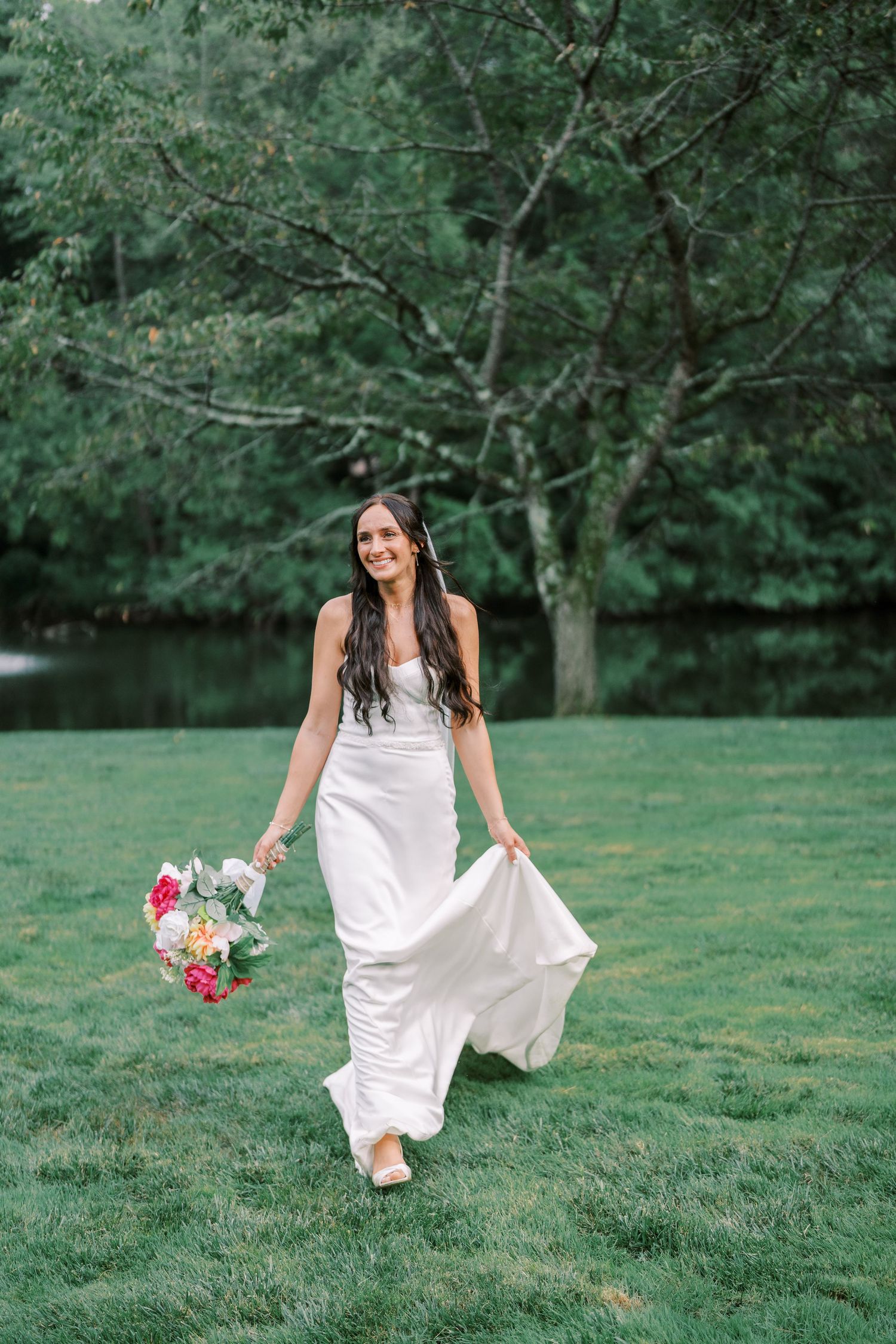 A bride in flowing white dress holds colorful bouquet while walking on grass near a pond.